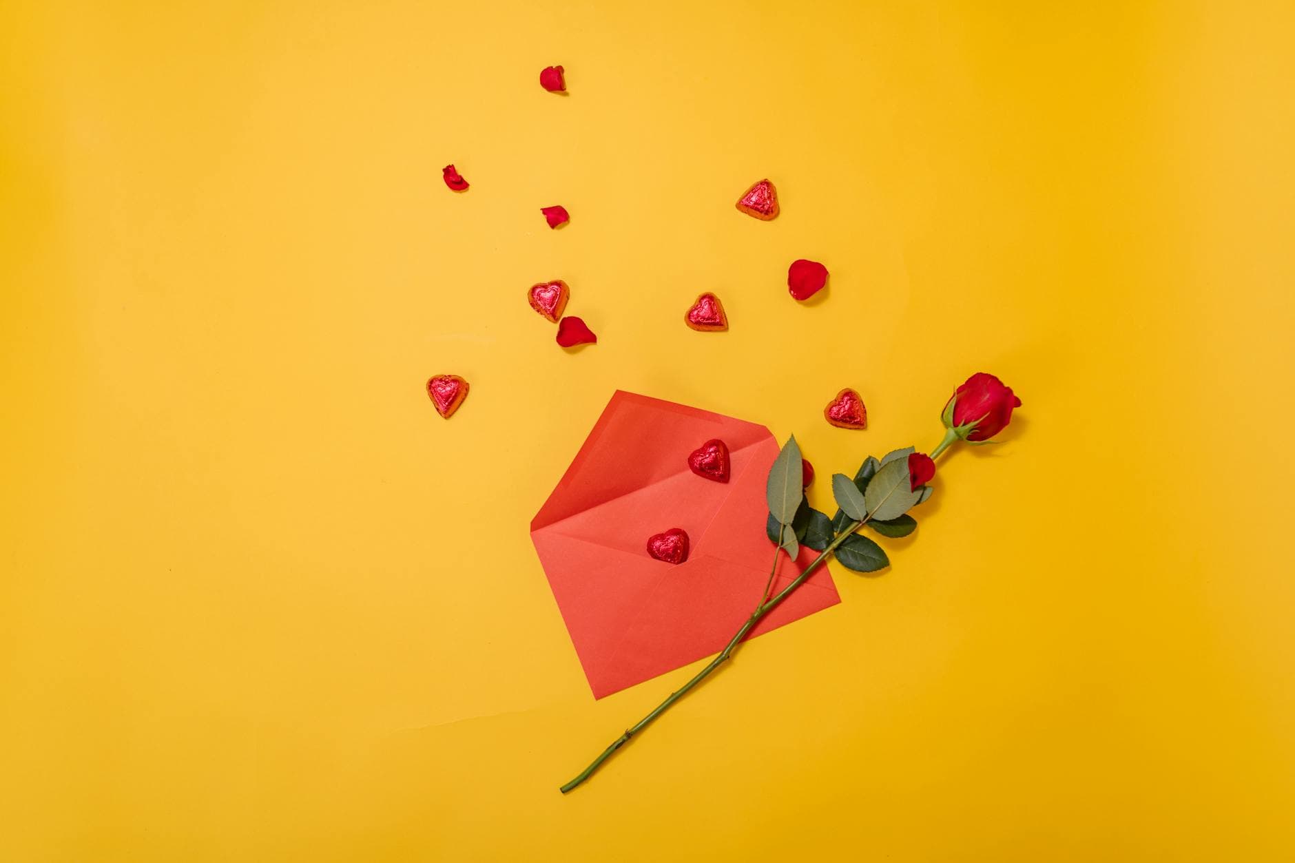 A romantic setting with a red rose, envelope, and heart-shaped candies on a yellow background. - valentine's day long distance ideas