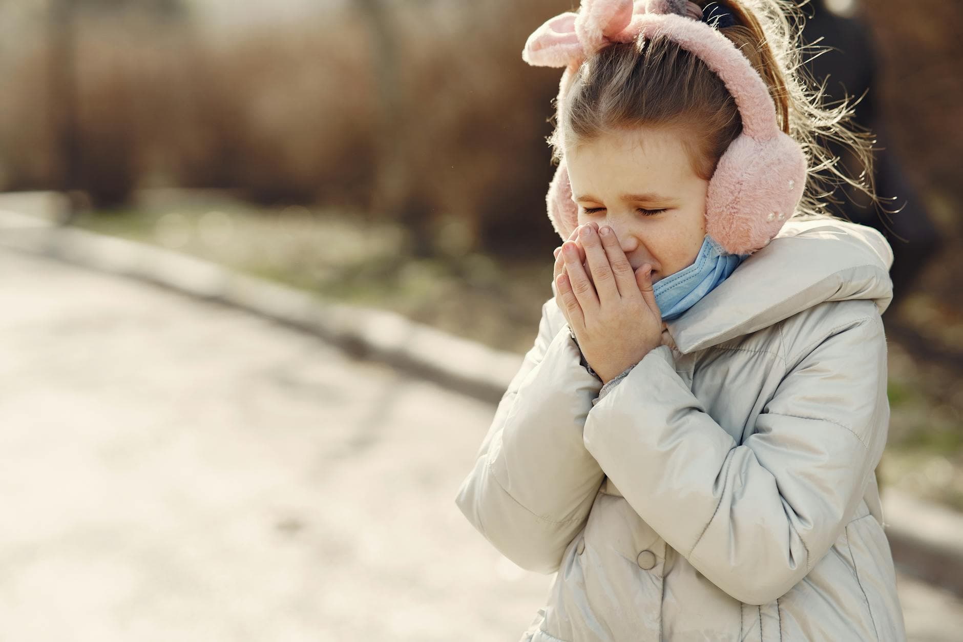 Small child with lowered face mask covering face by hands while sneezing on street - allergy relationship strain