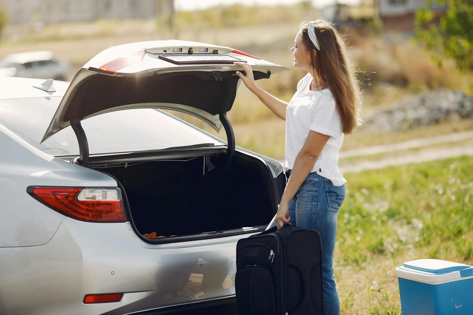 Side view of cheerful female traveler in headband and casual clothes putting luggage in open trunk of modern car while spending summer weekend in countryside - declutter emotional baggage