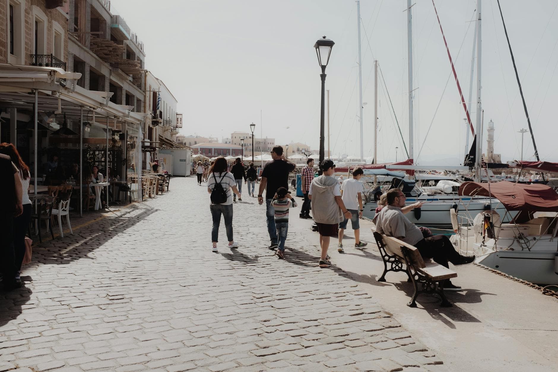 Tourists strolling along Chania harbor's cobblestone path, accompanied by docked boats under clear skies. - post holiday family talk