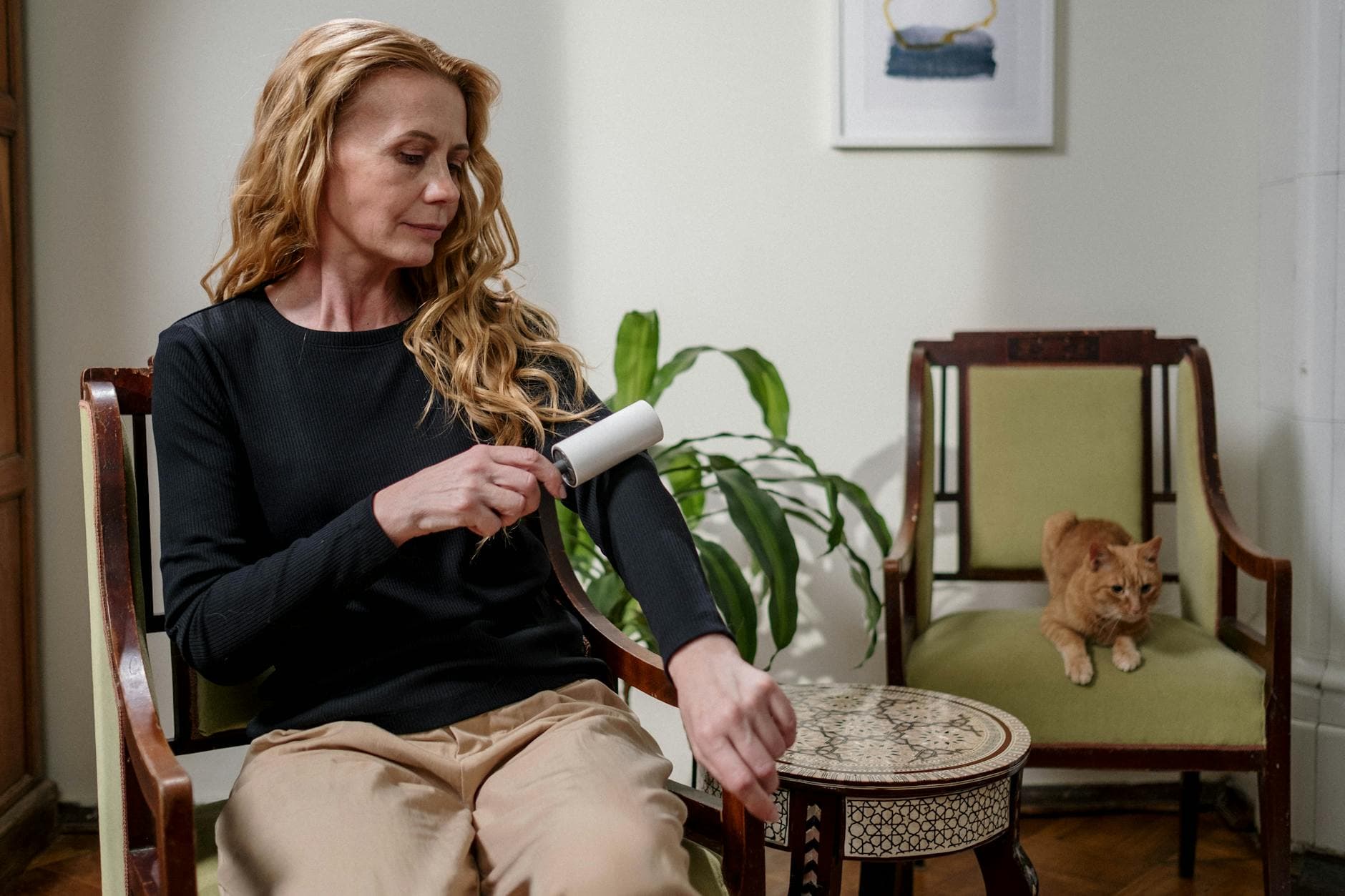 A woman cleaning her clothes with a lint roller while a ginger cat sits nearby, indoors. - spring allergy parenting
