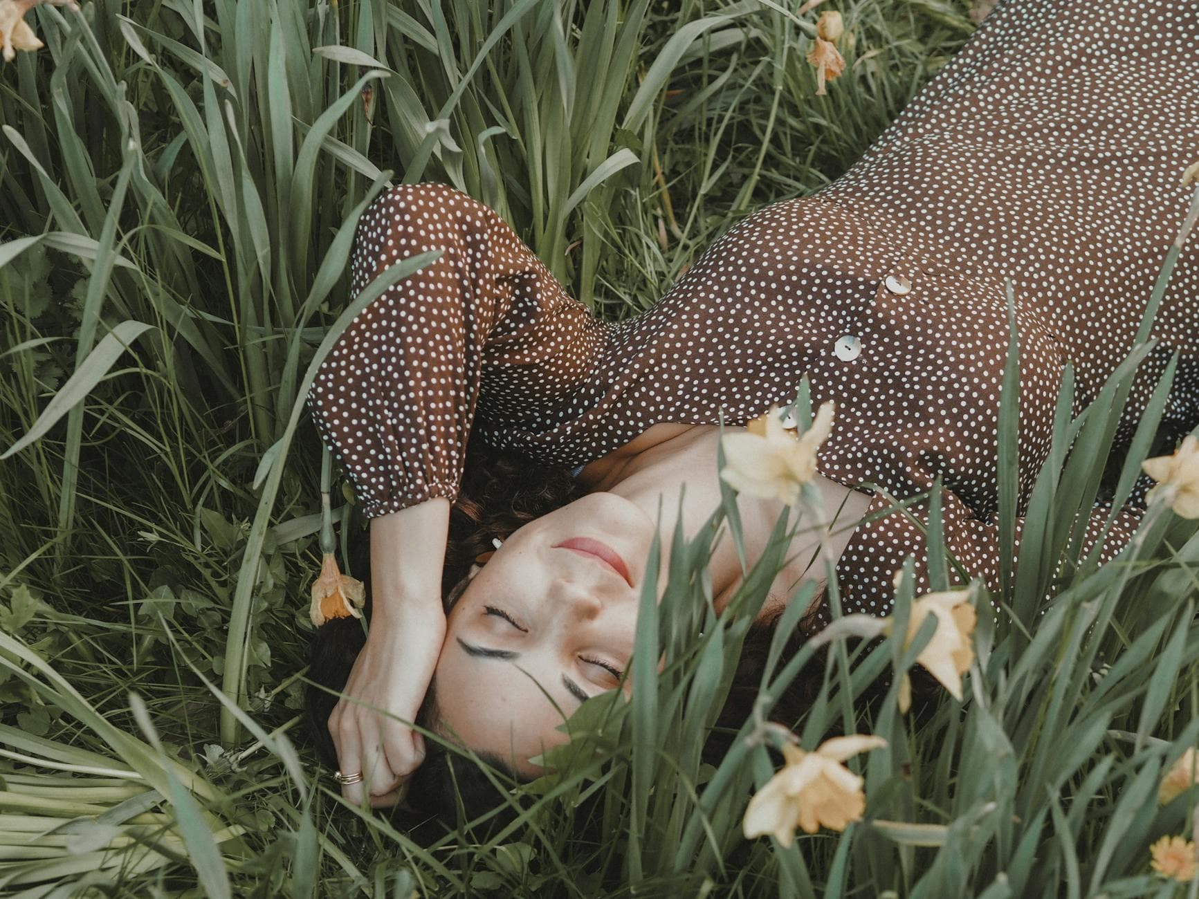 A serene woman in a polka dot dress rests among blooming flowers and green grass, embodying tranquility. - spring depression therapy