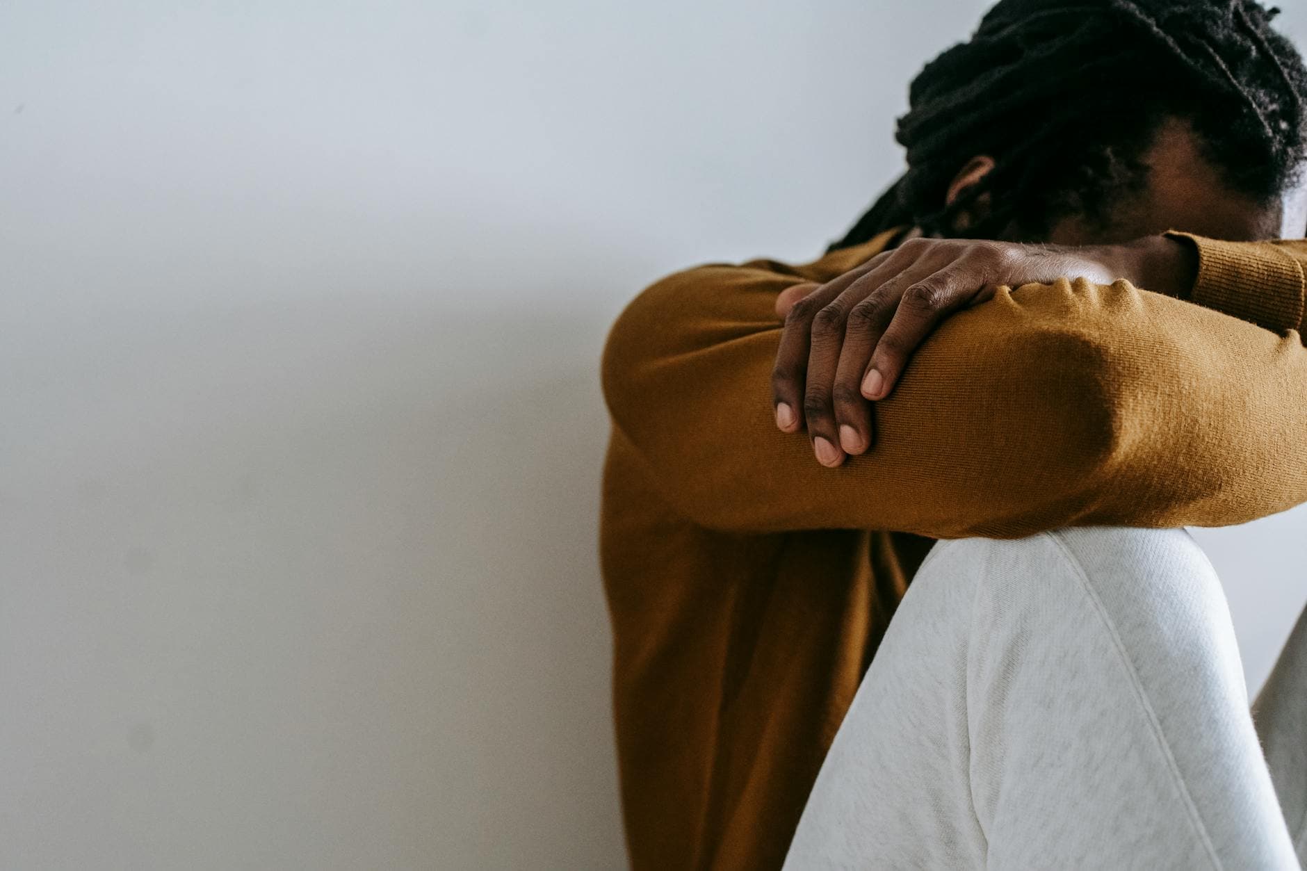 Crop anonymous depressed African American male embracing knees and covering face with arms while sitting on floor against white wall - spring overwhelm therapy