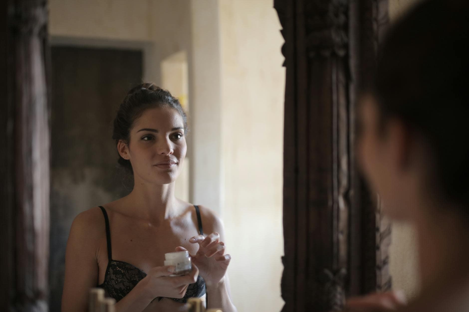 A woman in lingerie applies cream while looking at her reflection in a mirror. - spring self-care