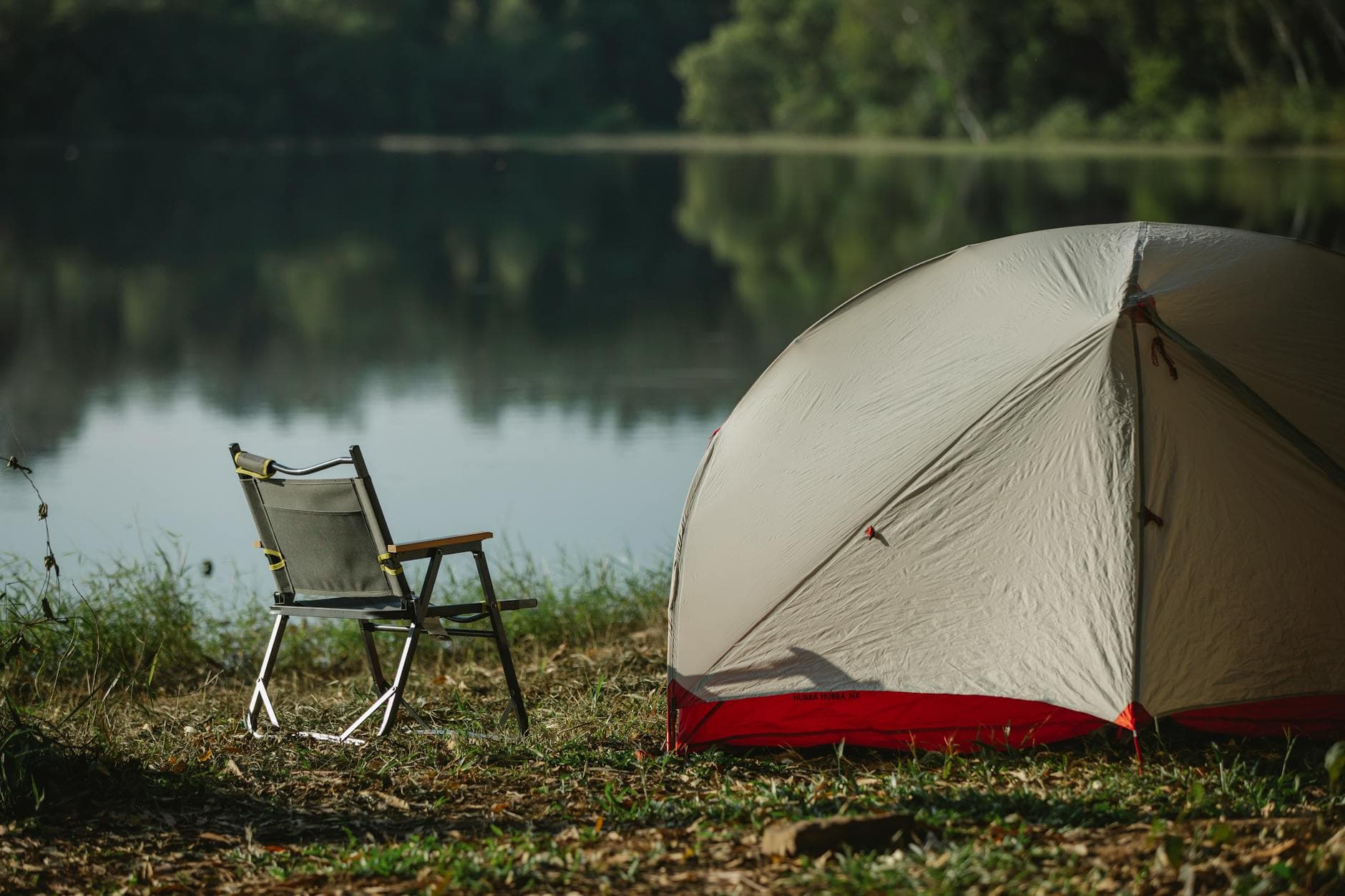 Tent and folding chair on grass coast against trees reflecting in lake in soft sunlight - summer camp transition