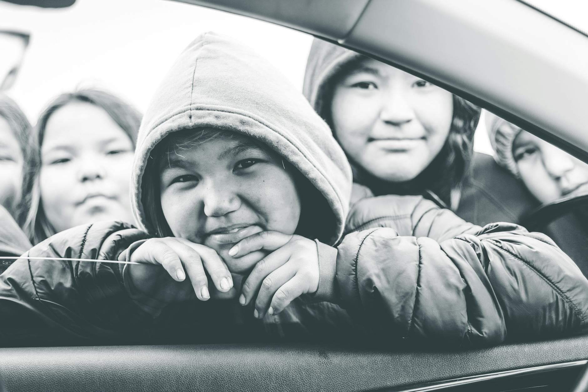 Group of children smiling and looking through a car window, wearing hooded jackets in a black and white image. - teen winter blues