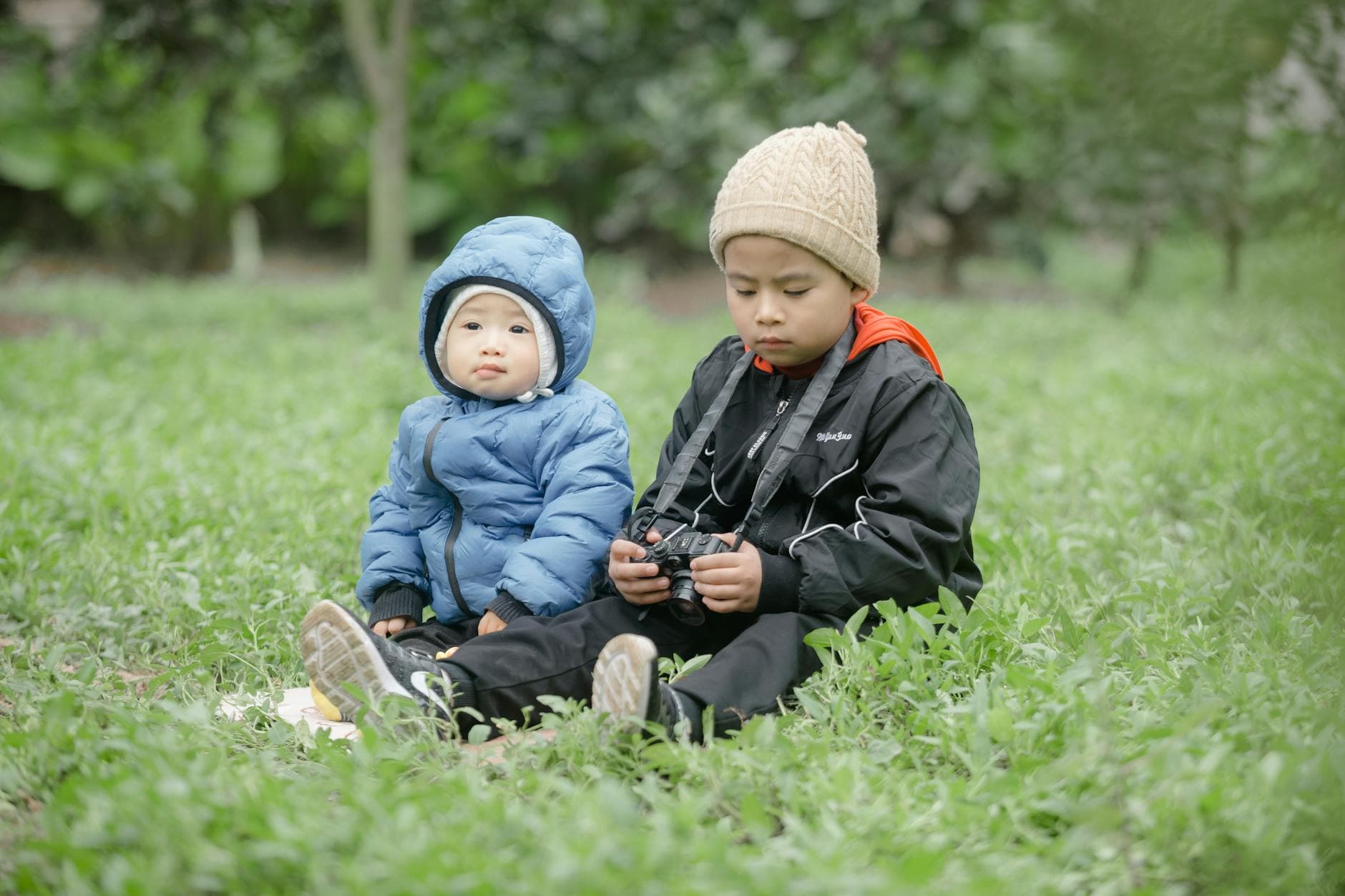 Two children sitting on grass in winter clothes, one holding a camera, in a tranquil outdoor setting. - toddler independence winter