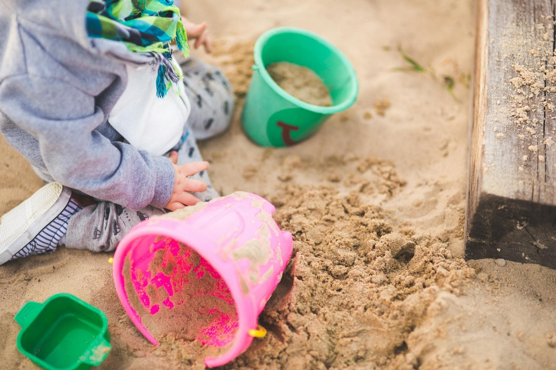 A child enjoys playing in a sandbox using vibrant colored buckets during a sunny day. - toddler outdoor play