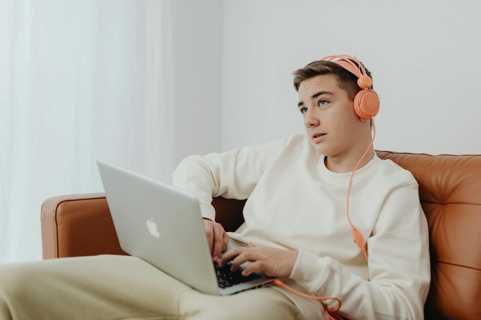 Teenage boy wearing headphones and using a laptop on a sofa, enjoying leisure time indoors. - active listening teen