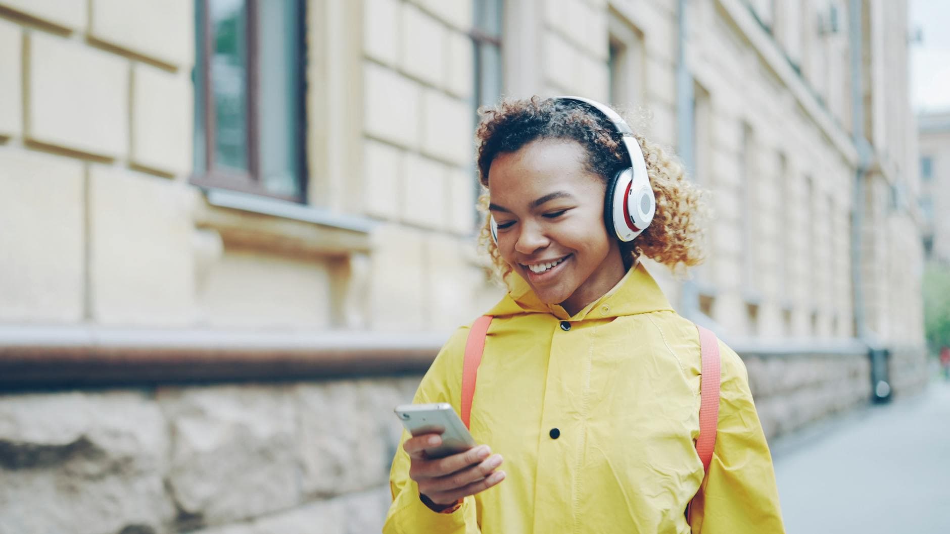 Smiling woman in yellow coat listening to music with headphones and smartphone outdoors. - active listening teens