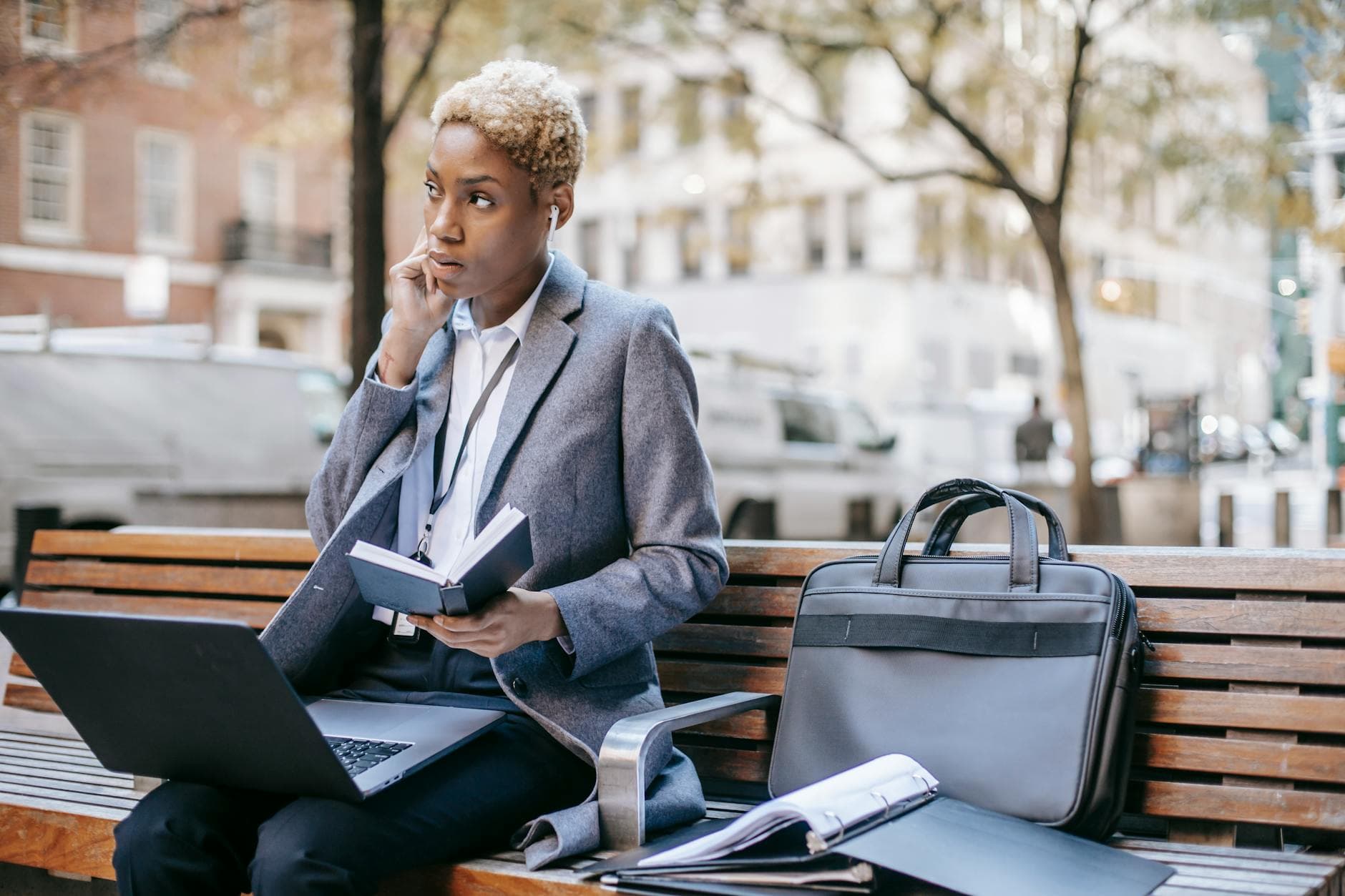 Serious young African American businesswoman with formal coat and earbuds sitting on bench with laptop on knees and doing paperwork in autumn city park - communicate needs busy season