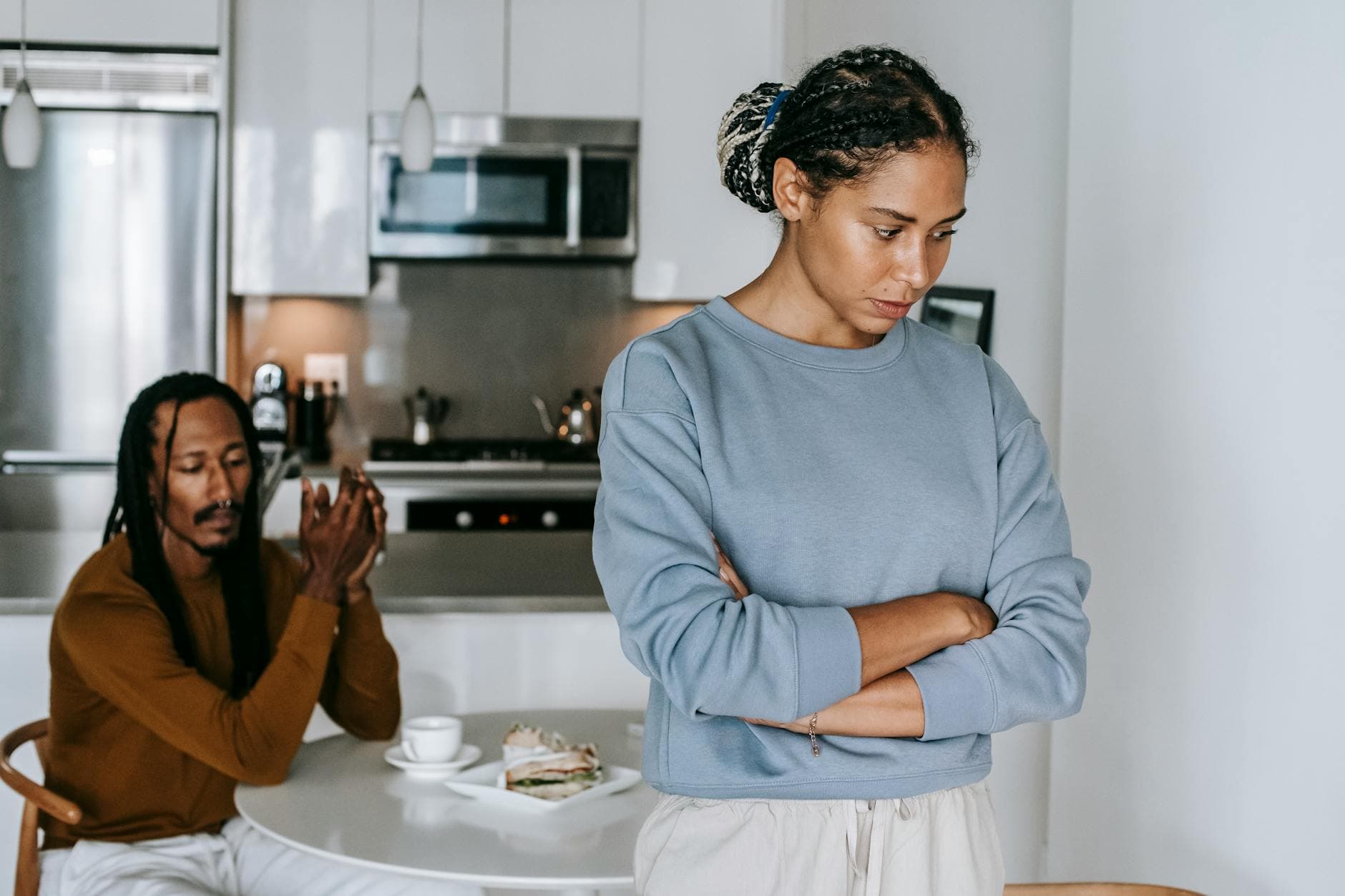 Young discontented African American female with folded arms against male partner at kitchen table during quarrel - couple conflict resolution