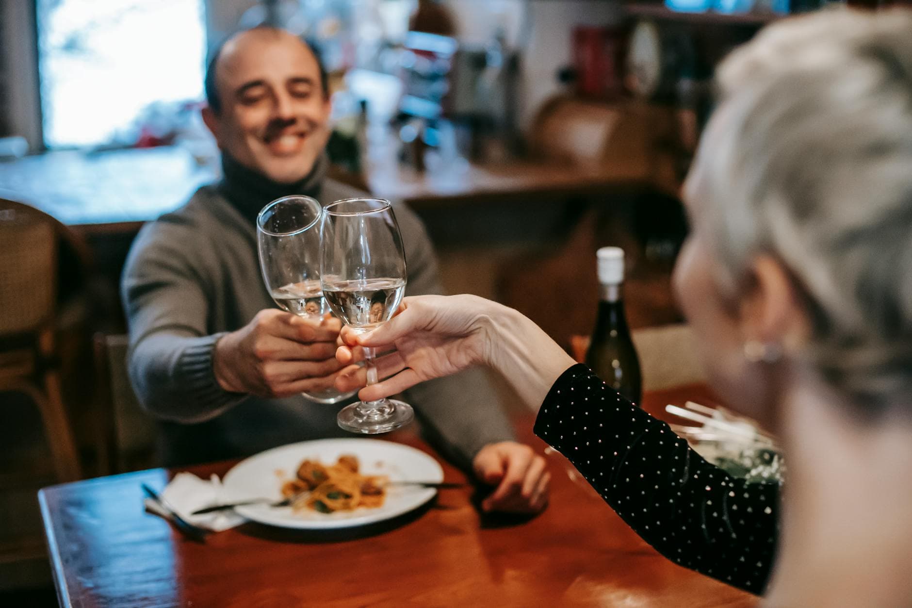 Cheerful adult couple in casual outfits clinking glasses with white wine while having dinner in restaurant - dating over 40