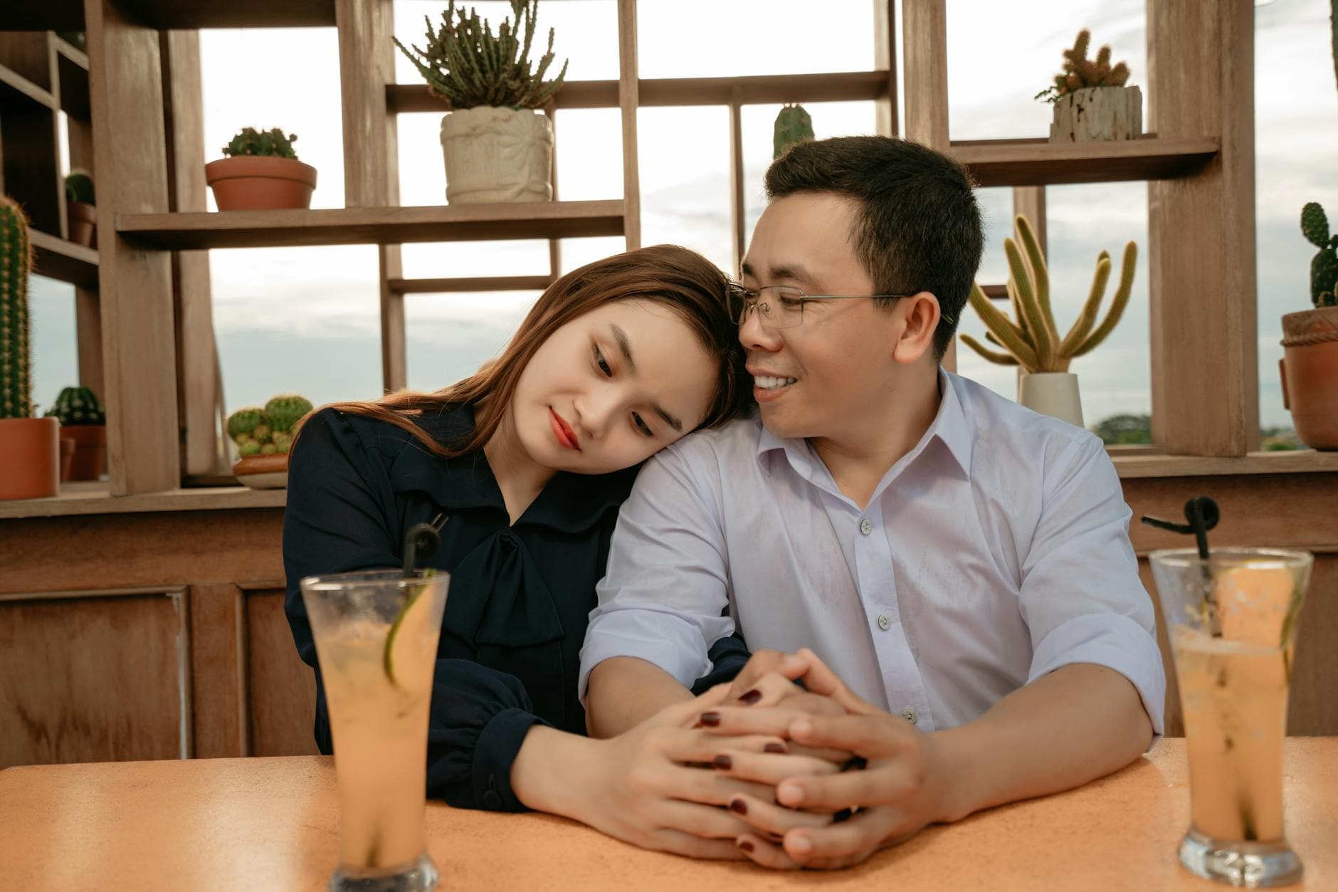 Positive ethnic couple in elegant outfits sitting at table with cocktails on date in cafe and holding hands while hugging in daylight with potted plants on shelves on background - dating in 40s