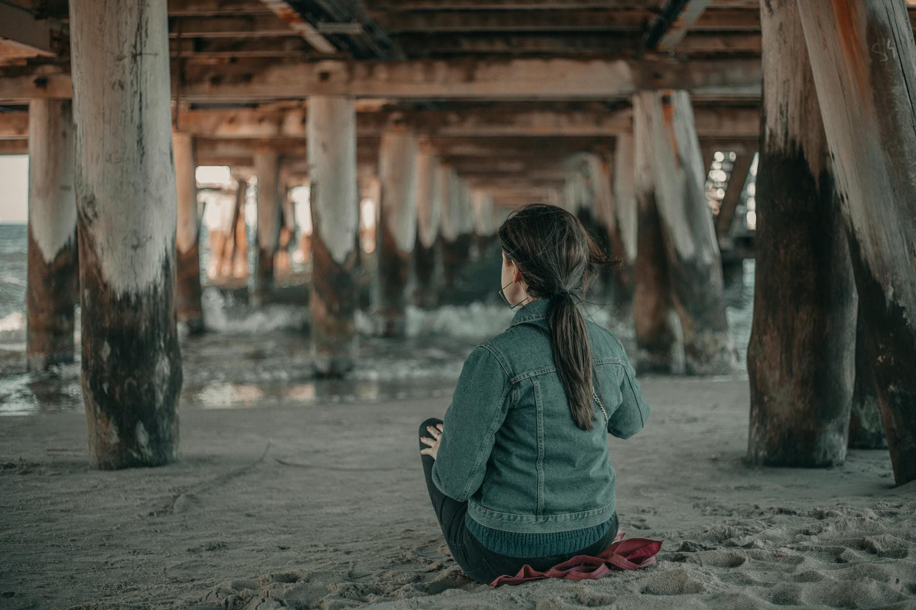 Back view of anonymous female tourist sitting on sandy coast near rippling sea under shabby wooden bridge in coastal terrain - letting go resentment