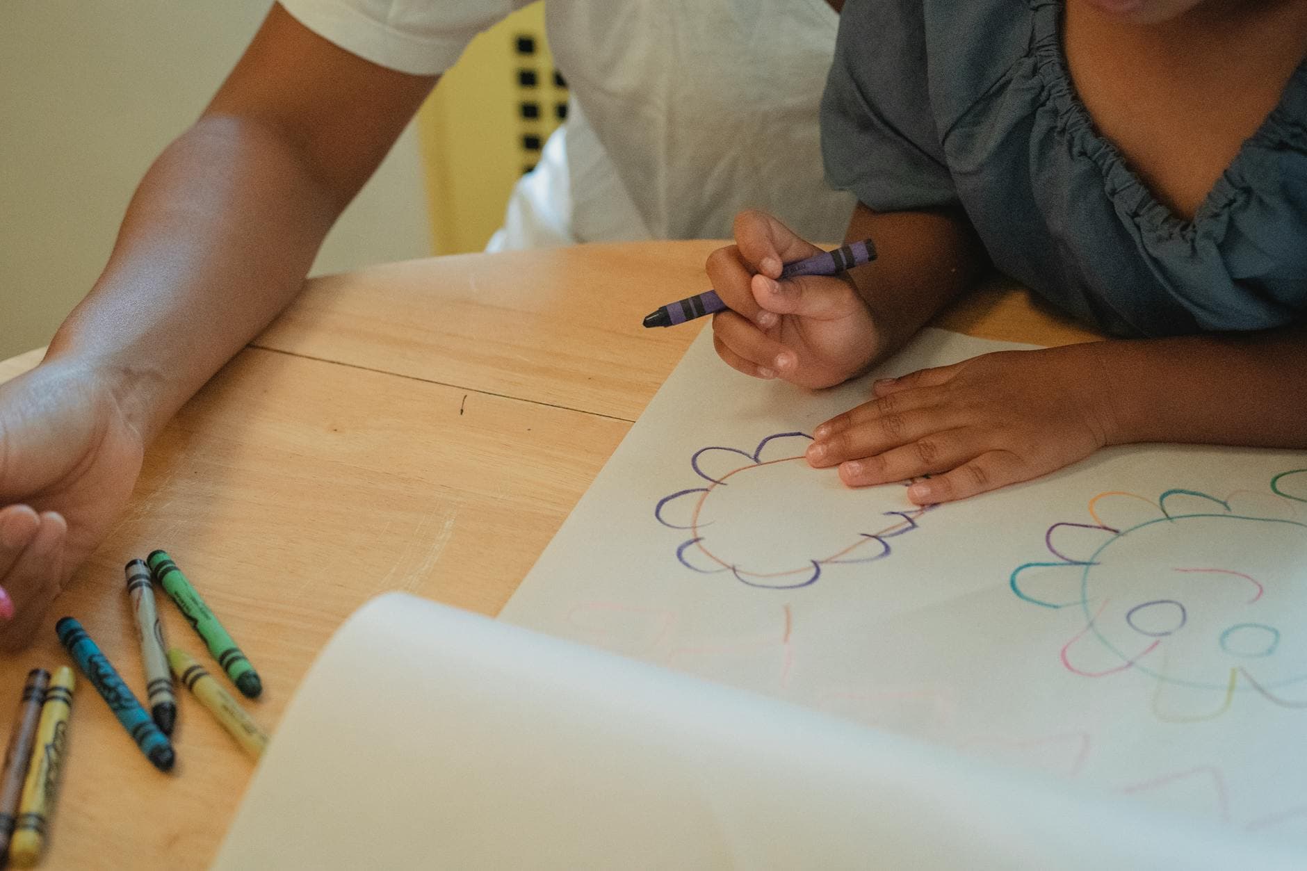 Crop anonymous mother and daughter sitting at wooden table and drawing with color wax crayons - preschool spring activities
