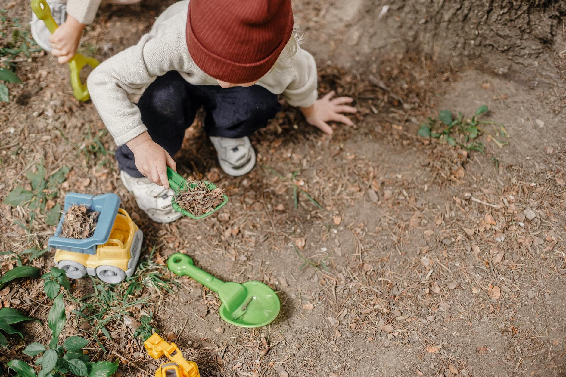 High angle of little kid in brown hat squatting on ground and playing with plastic colourful toys - preschool spring changes