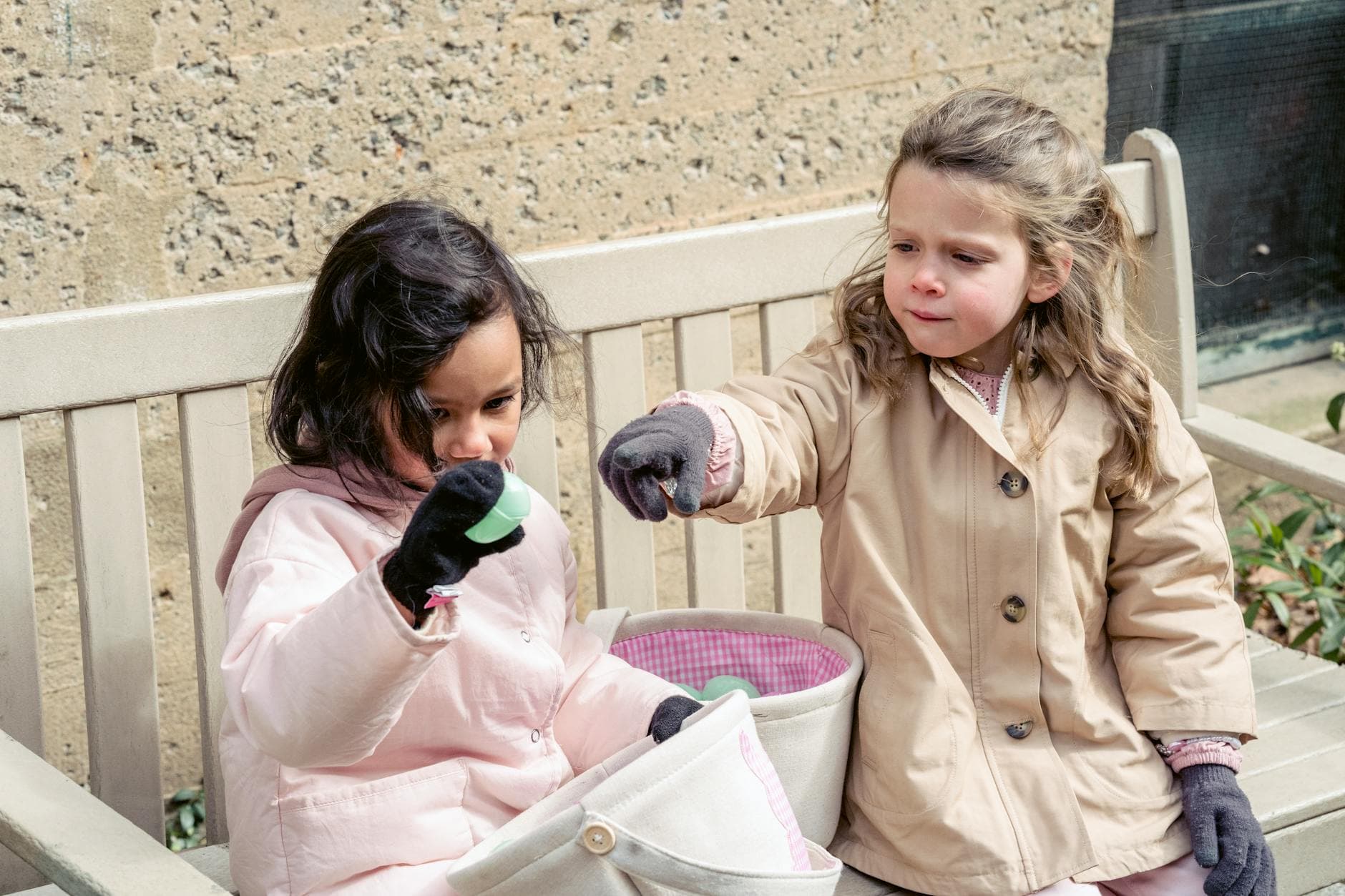Multiethnic girls wearing warm clothes playing with colorful Easter eggs on bench in sunny spring park - preschooler spring play