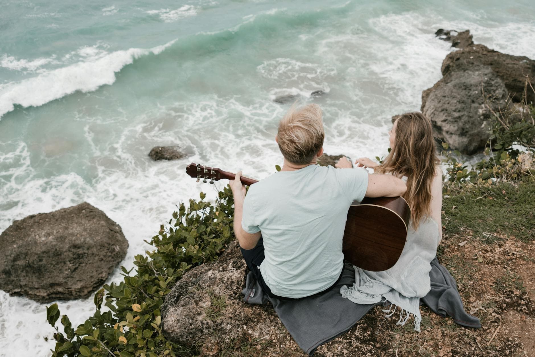 From above back view of anonymous romantic travelers playing acoustic guitar sitting together on rock above waving ocean during date - reconnect with partner