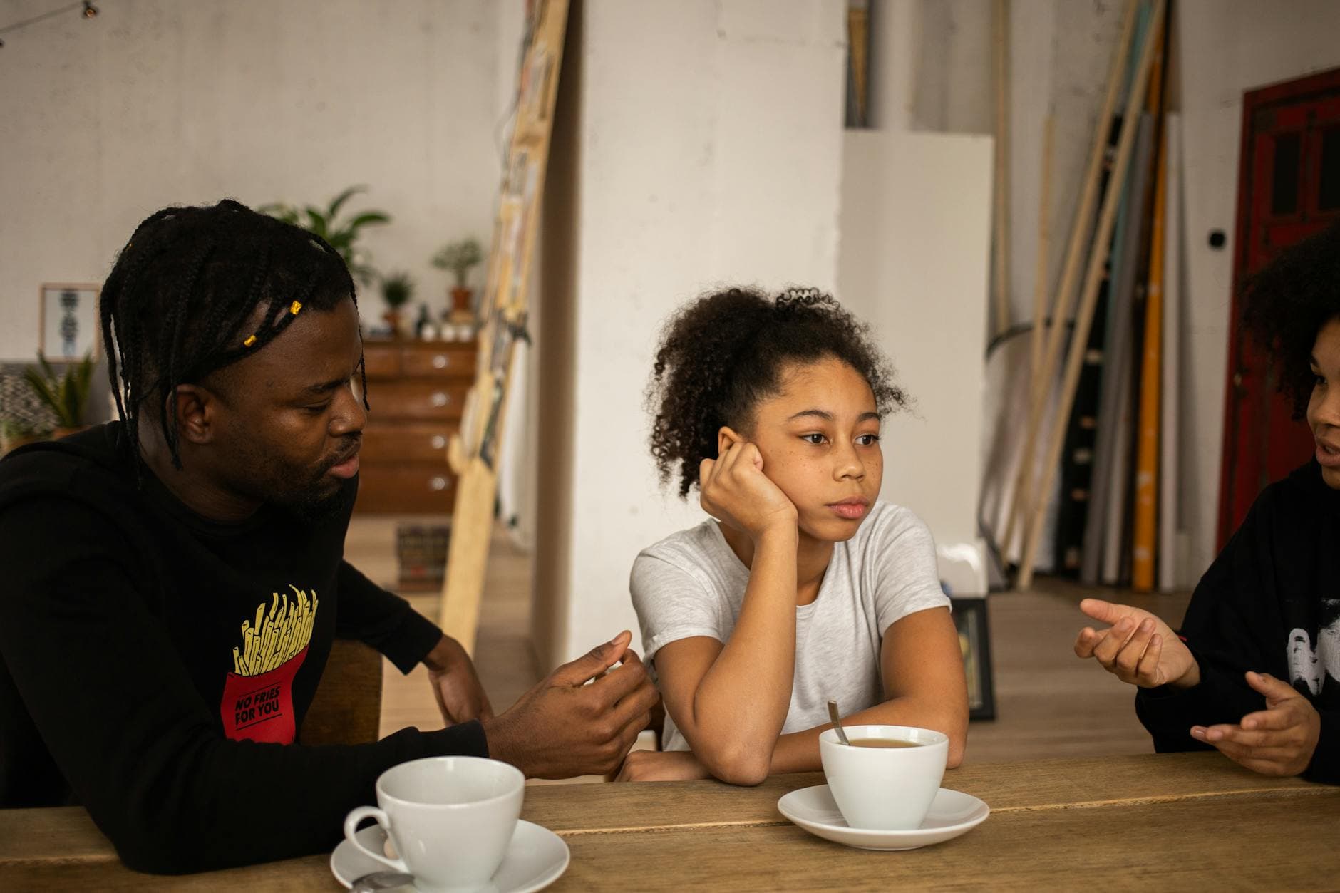 Unhappy African American girl sitting at table with cups of coffee between faceless mother and father having quarrel in room - active listening family