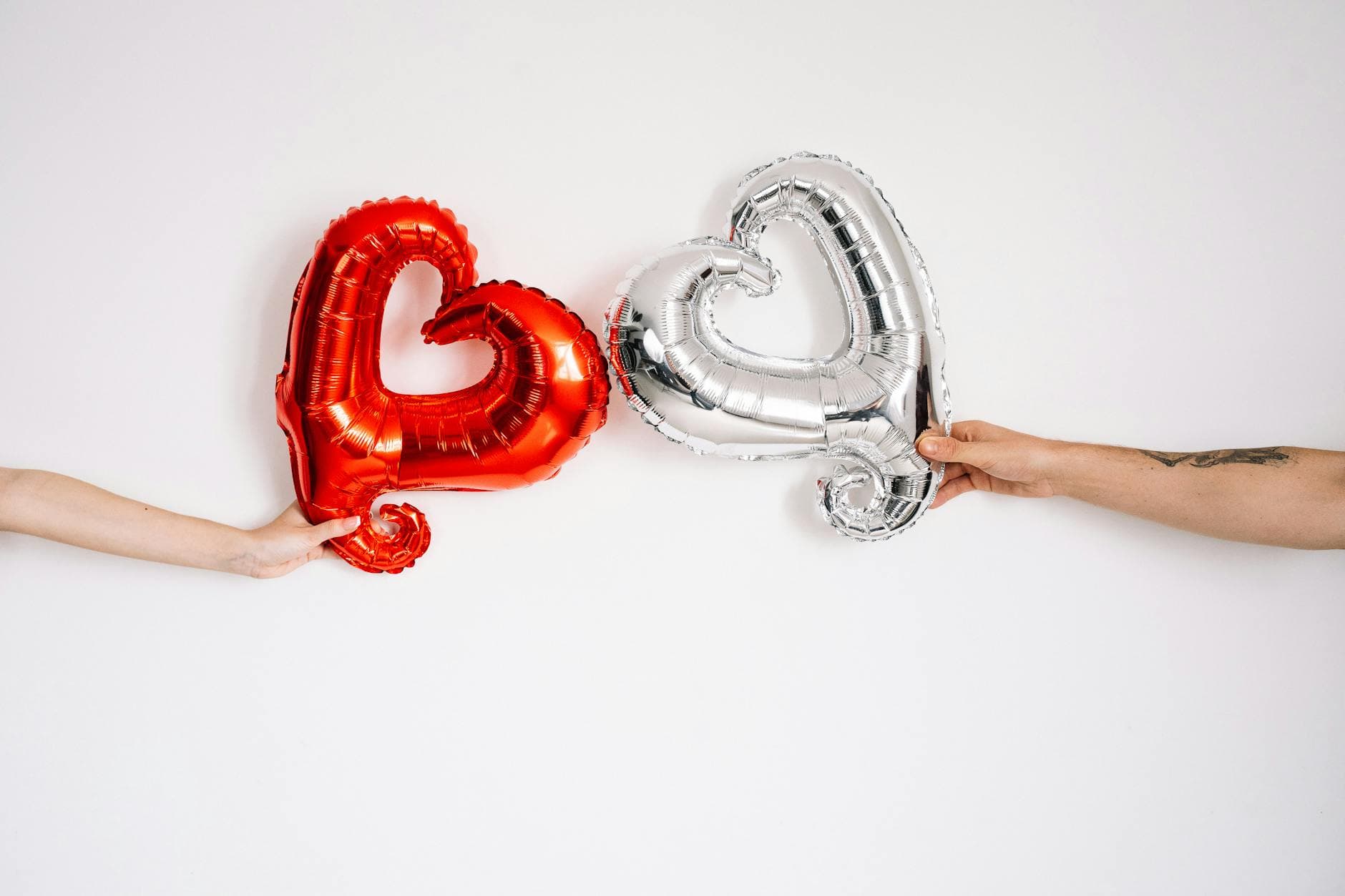 Two hands hold red and silver heart-shaped balloons against a white background. - long distance date ideas