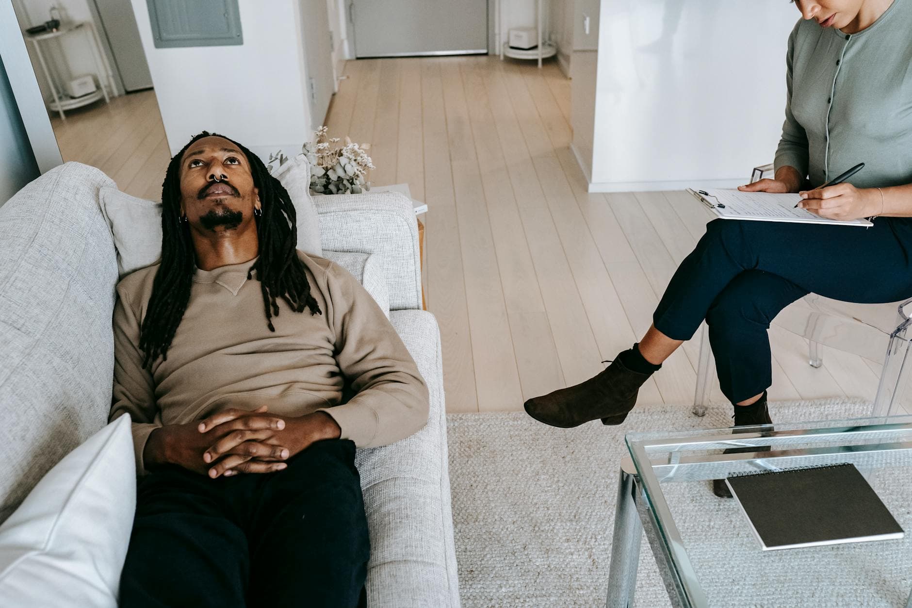 From above of African American male looking up while lying on couch near crop female therapist writing notes on clipboard during session - mental health habits