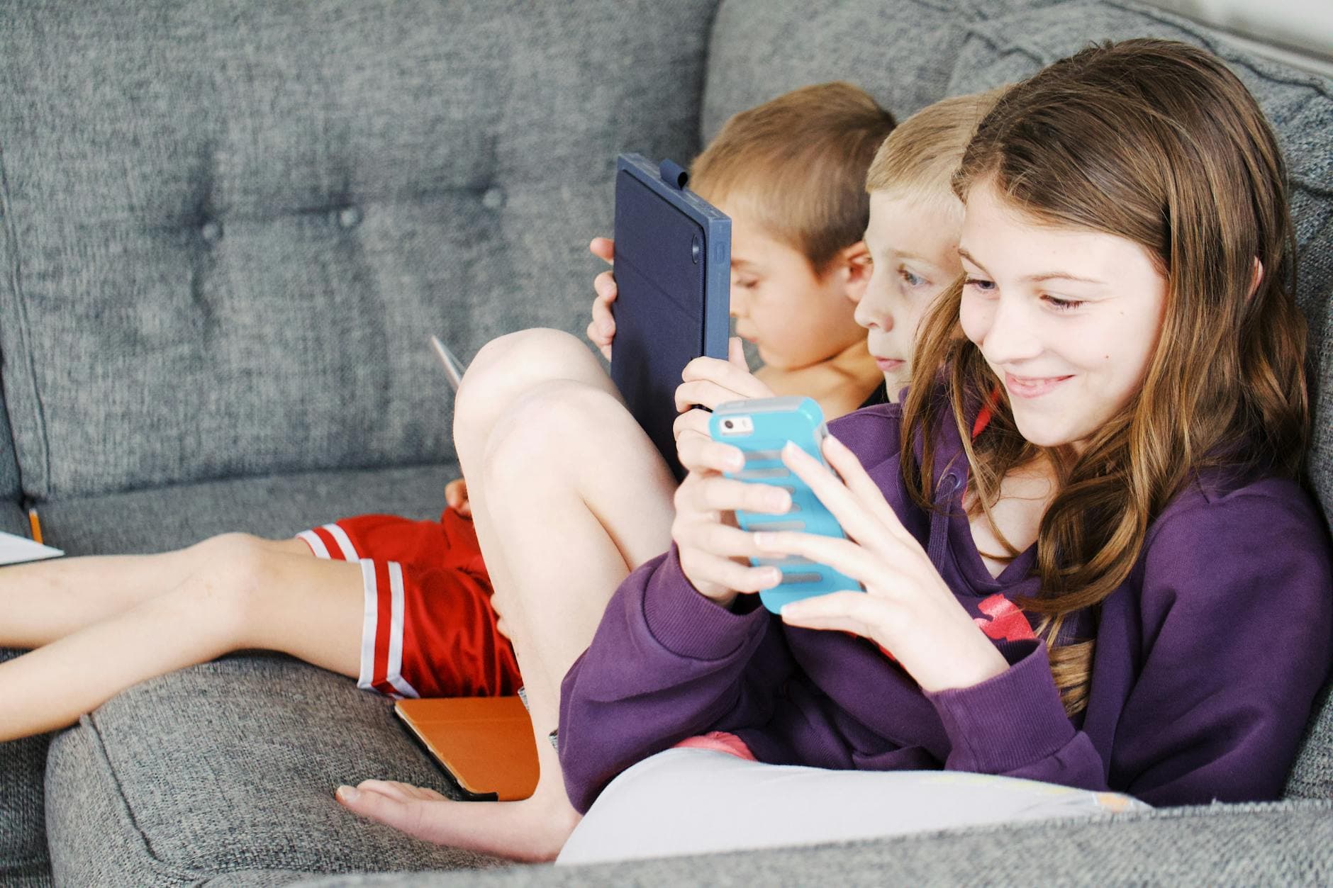 Three children sitting on a couch, engaged with electronic devices, smiling and enjoying leisure time. - screen time limits