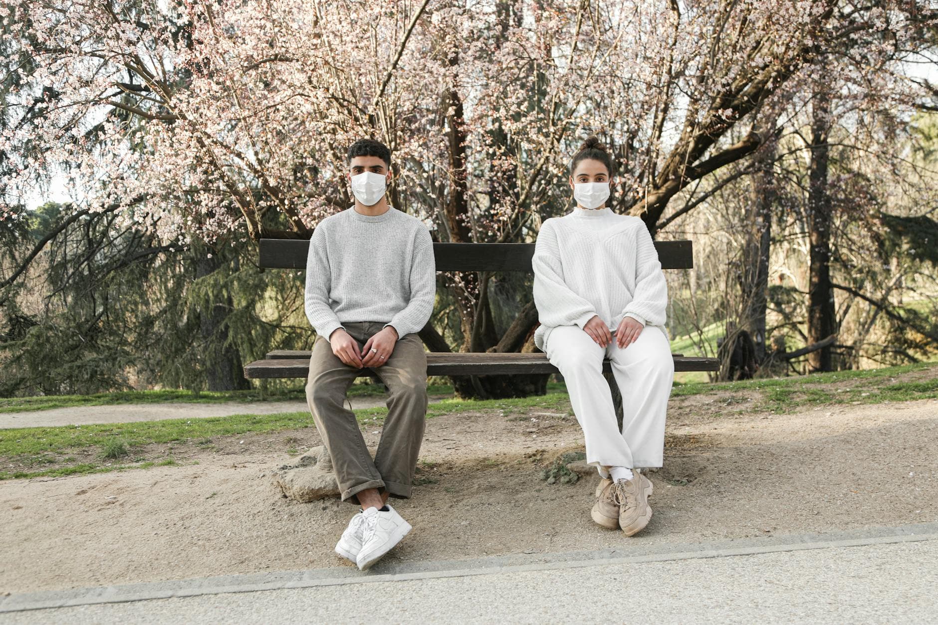 A man and woman in masks sit apart on a bench under blooming trees, reflecting social distancing. - spring outdoor safety