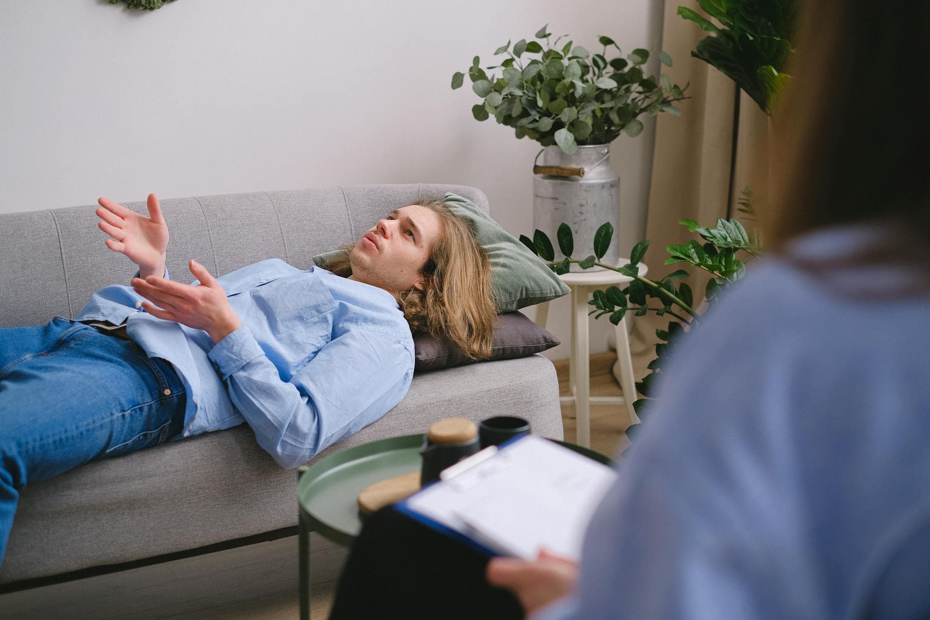 Concerned male client lying on sofa and speaking about mental problems with unrecognizable psychologist with clipboard during psychotherapy appointment in office - cbt for holiday anxiety