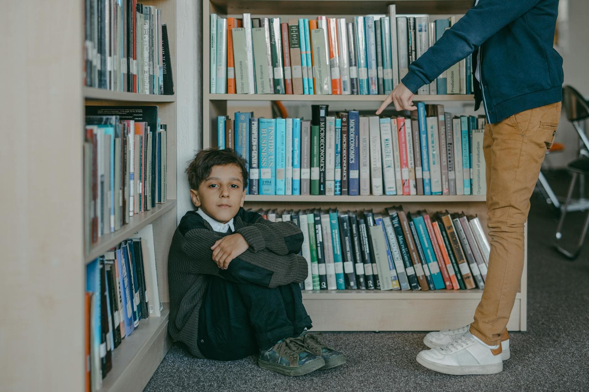 A sad boy sitting on the library floor while being bullied by a peer. - child social emotional development break