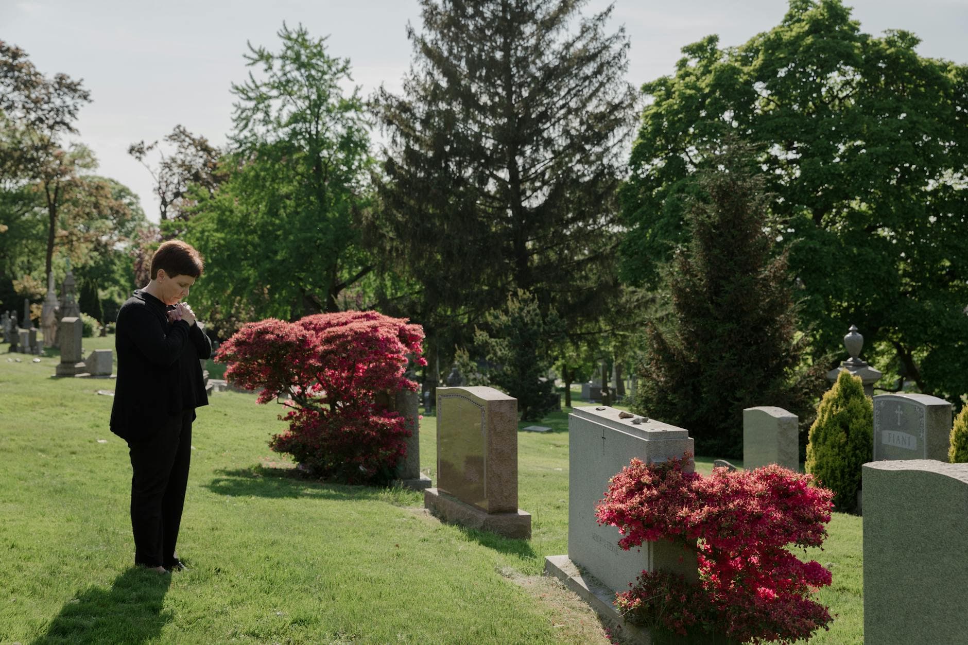 A woman stands thoughtfully in a picturesque cemetery, surrounded by green trees and vibrant flowers. - coping with grief in winter