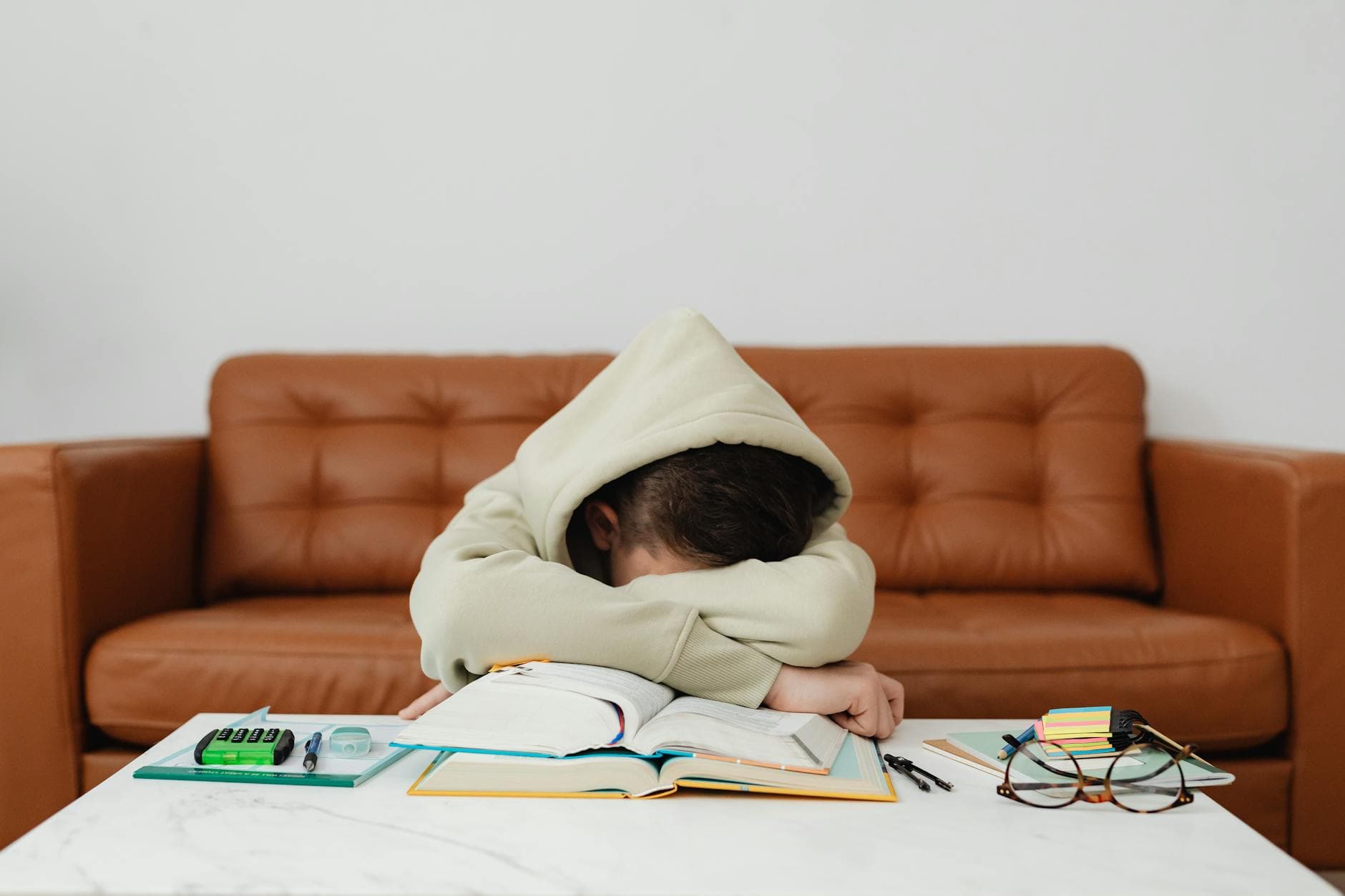 Child in hoodie leans over books, exhausted while studying on a couch. - kids winter break boredom