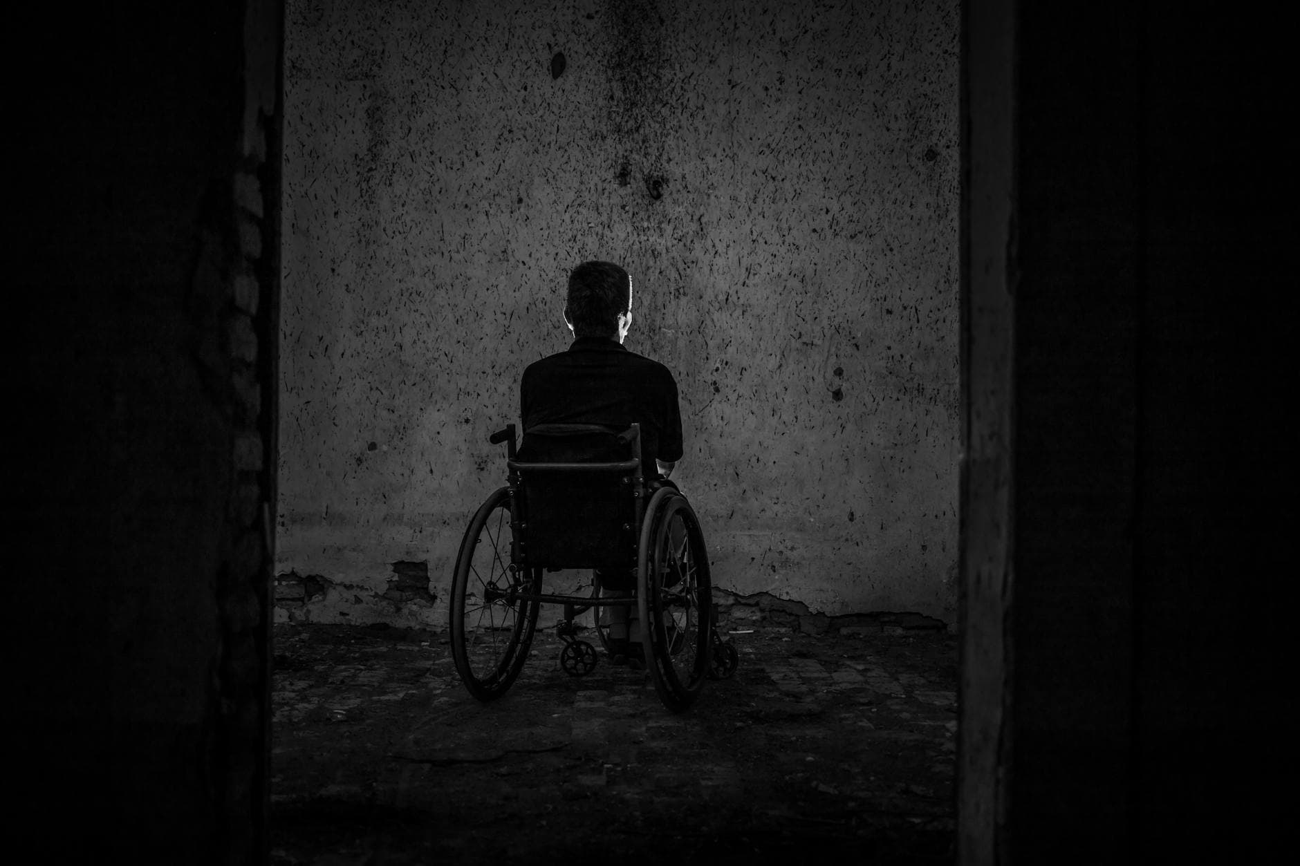 A man sits alone in a wheelchair facing a wall in a dark, empty room, evoking solitude. - overcoming winter loneliness