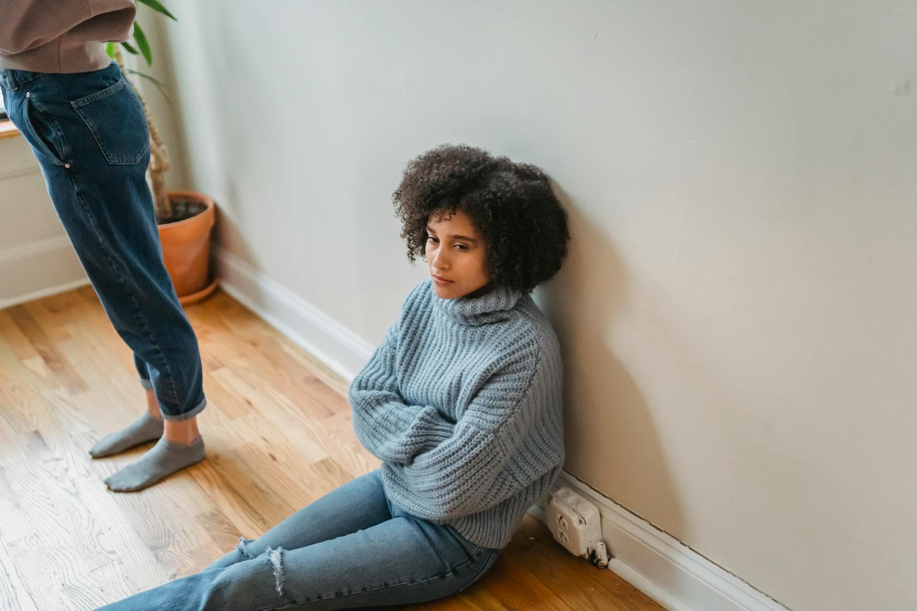 A young woman with curly hair sits thoughtfully by a wall indoors, wearing a blue sweater. - avoidant attachment style