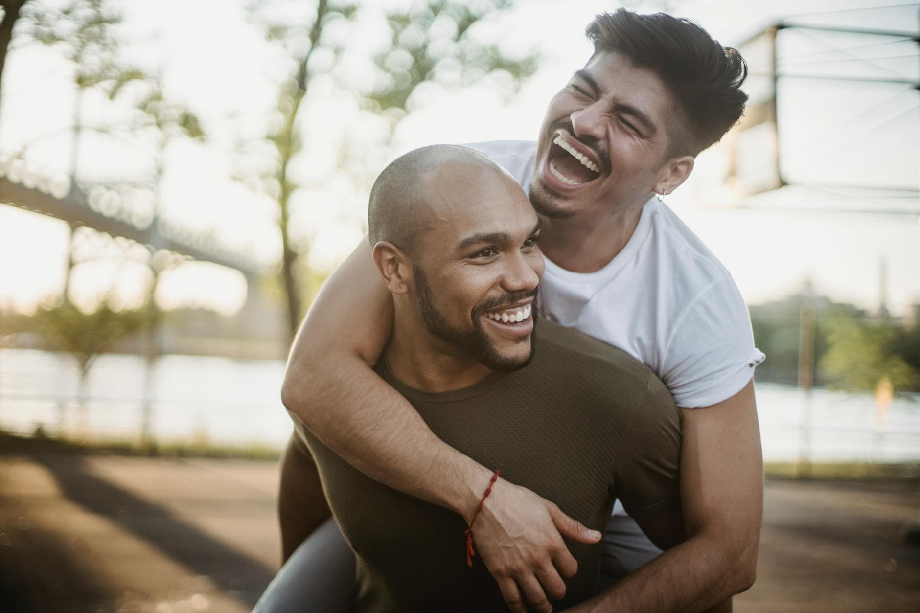 Happy interracial gay couple enjoying a playful piggyback ride in a sunny park setting. - express needs couples