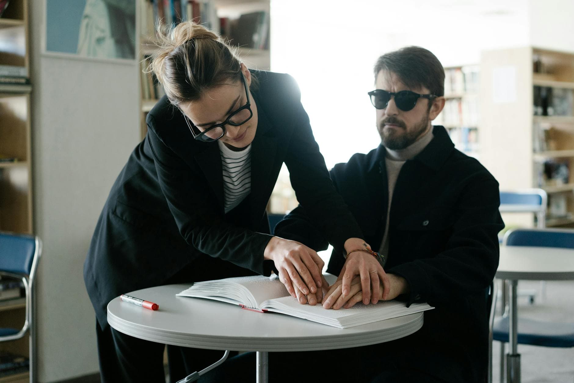 A caregiver assists a blind man in reading Braille at a library table. - self-compassion guide