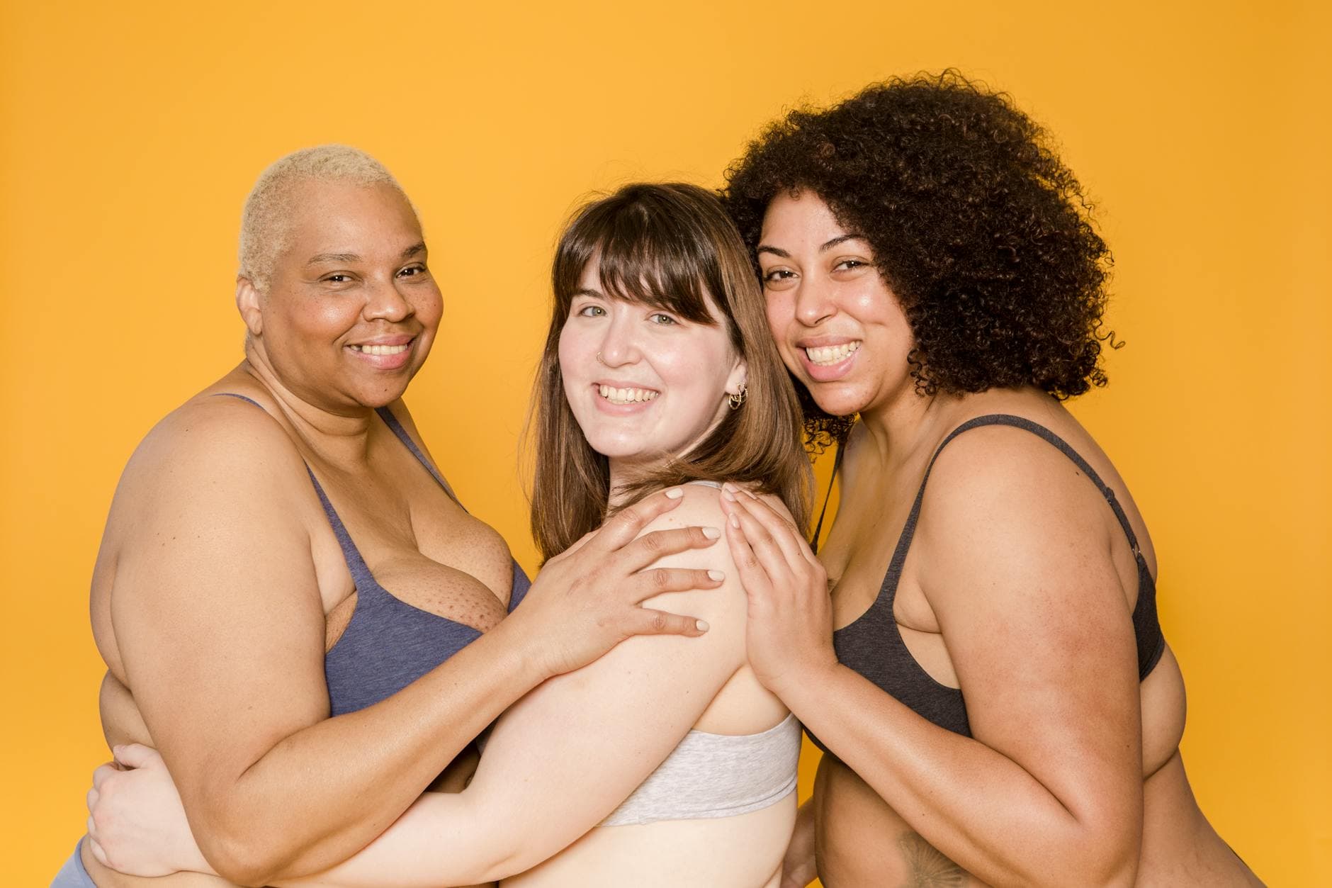 Three diverse women embracing confidently, promoting body positivity against a yellow background. - self compassion spring