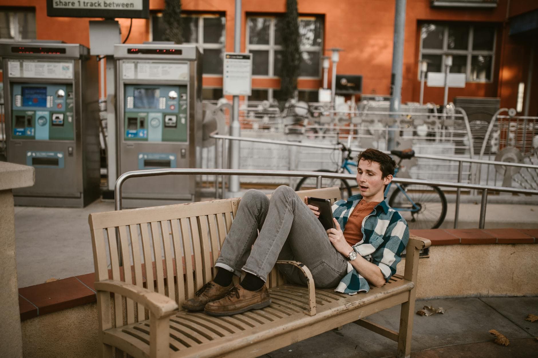 A young man reclining on a bench outdoors while using a smartphone near a train station. - spring relationship goals