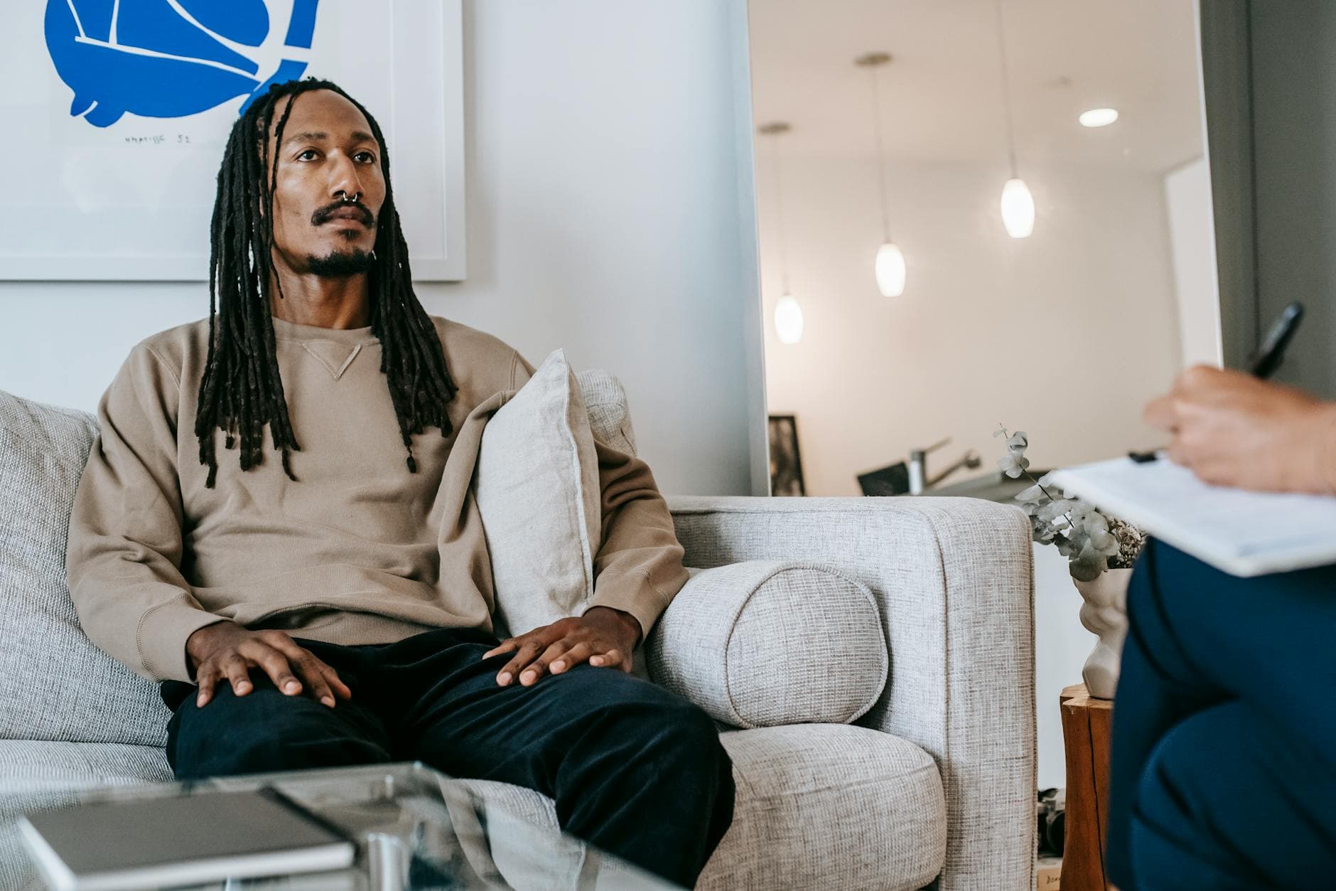 Pensive African American male client with dreadlocks sitting on couch with hands on laps near crop psychologist with clipboard during appointment - therapist for depression