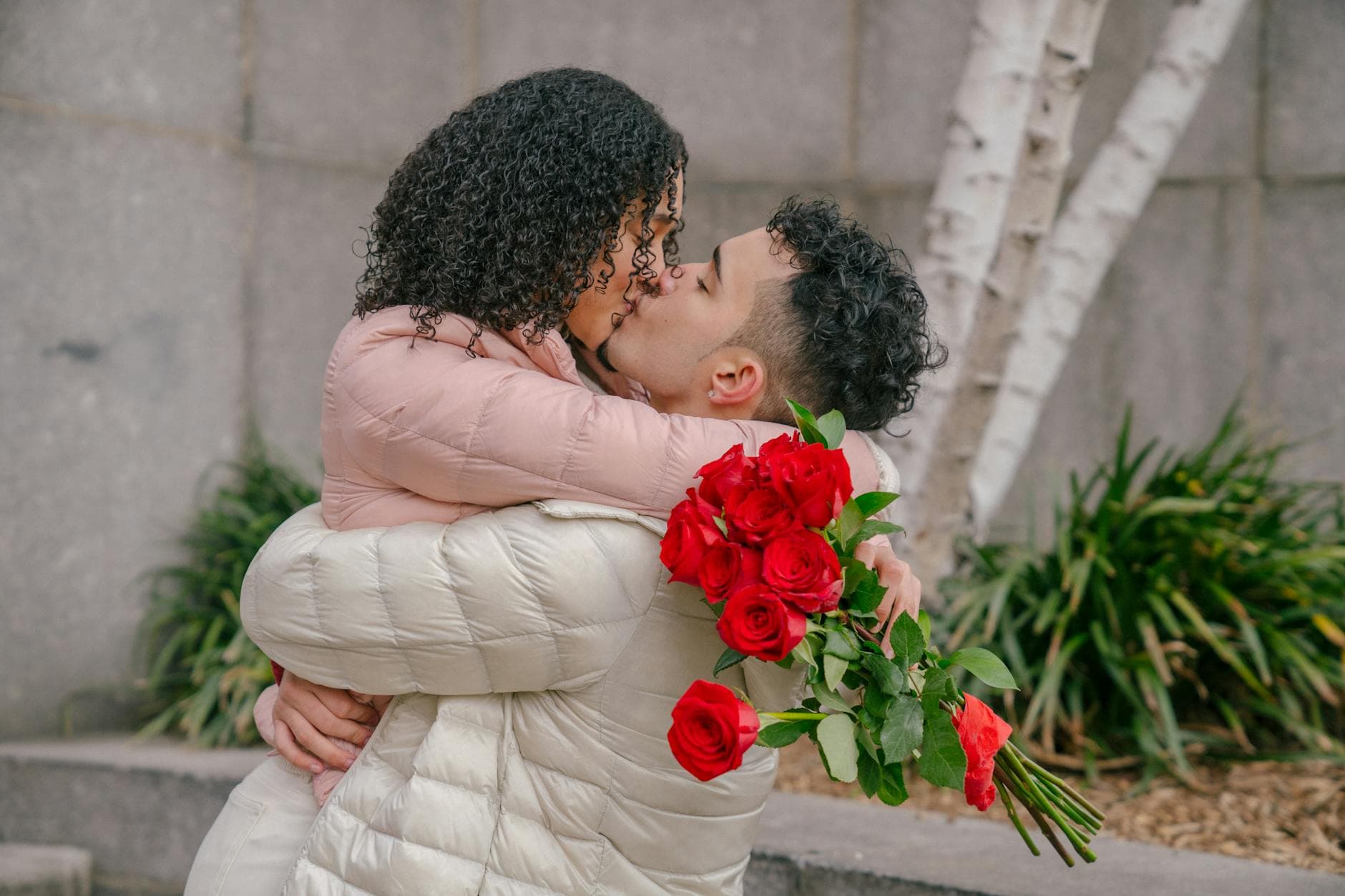 Side view of enamored young ethnic guy with curly hair hugging and kissing girlfriend after giving elegant bouquet of fresh red roses in park - cheap spring dates