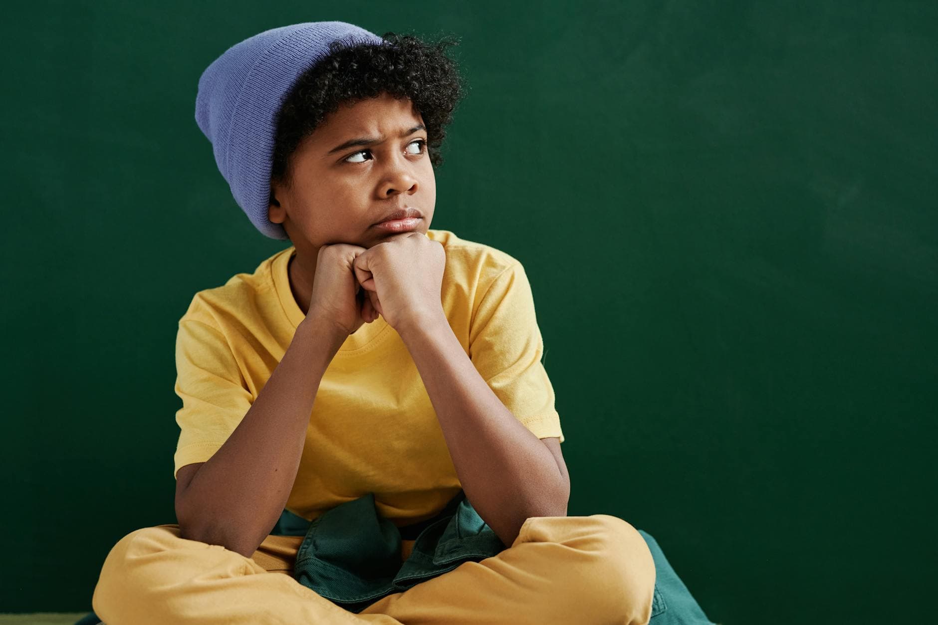 Pensive boy with curly hair wearing a yellow shirt and blue beanie against a green background. - child emotional intelligence