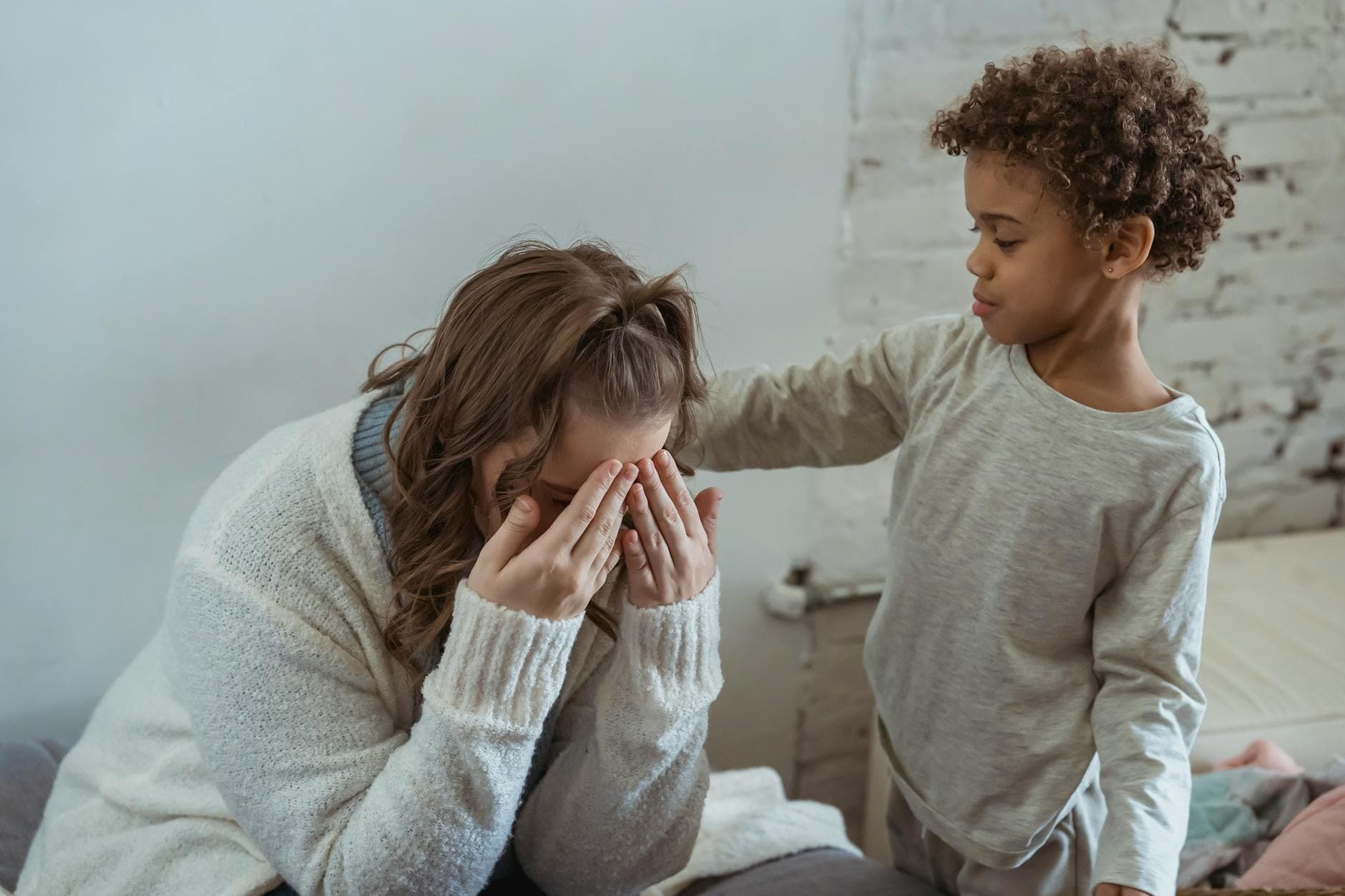 Caring African American son touching shoulder of upset faceless mother covering face while sitting in light room near wall at home - kids cope change