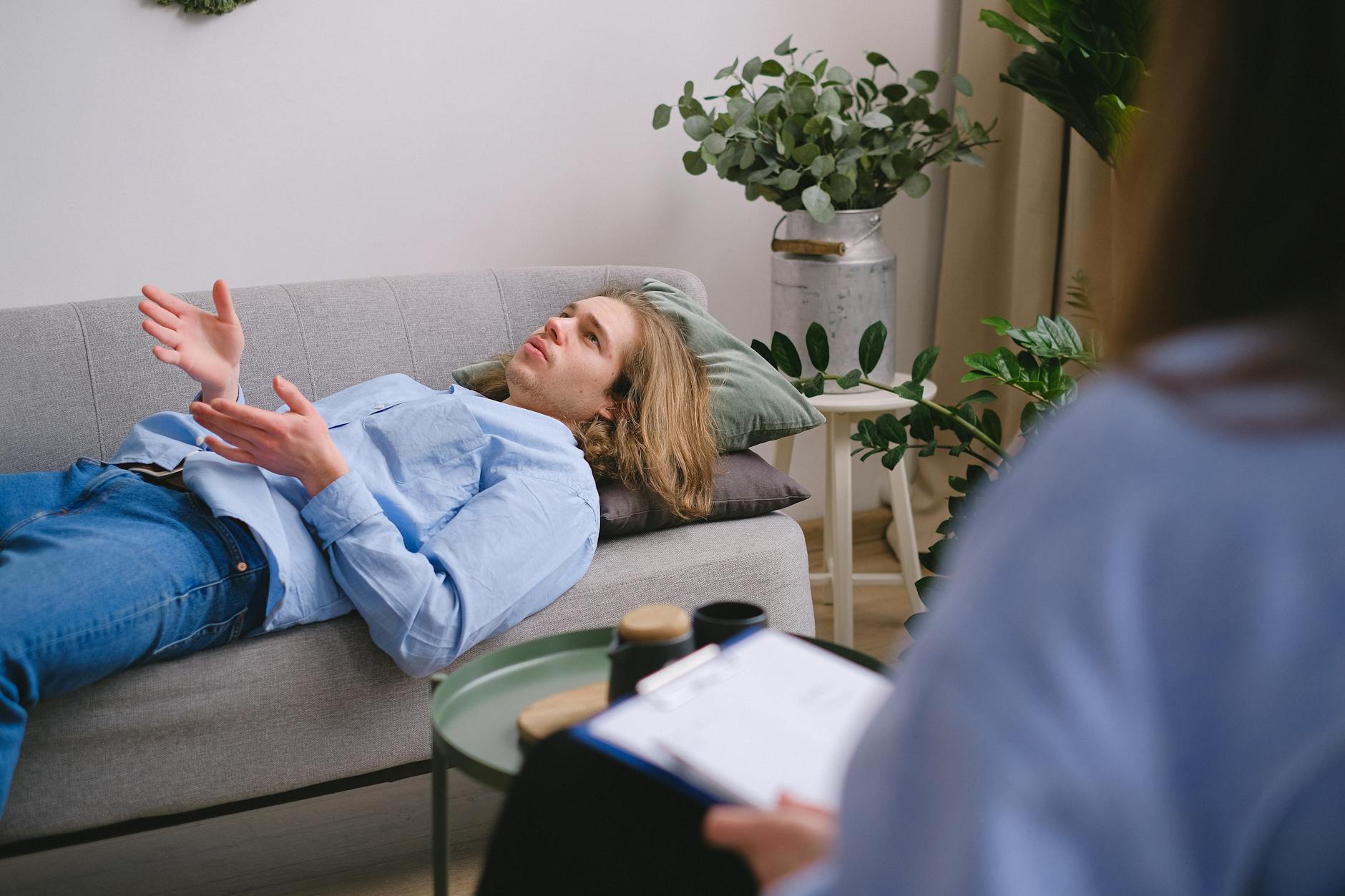 Concerned male client lying on sofa and speaking about mental problems with unrecognizable psychologist with clipboard during psychotherapy appointment in office - cbt for winter depression