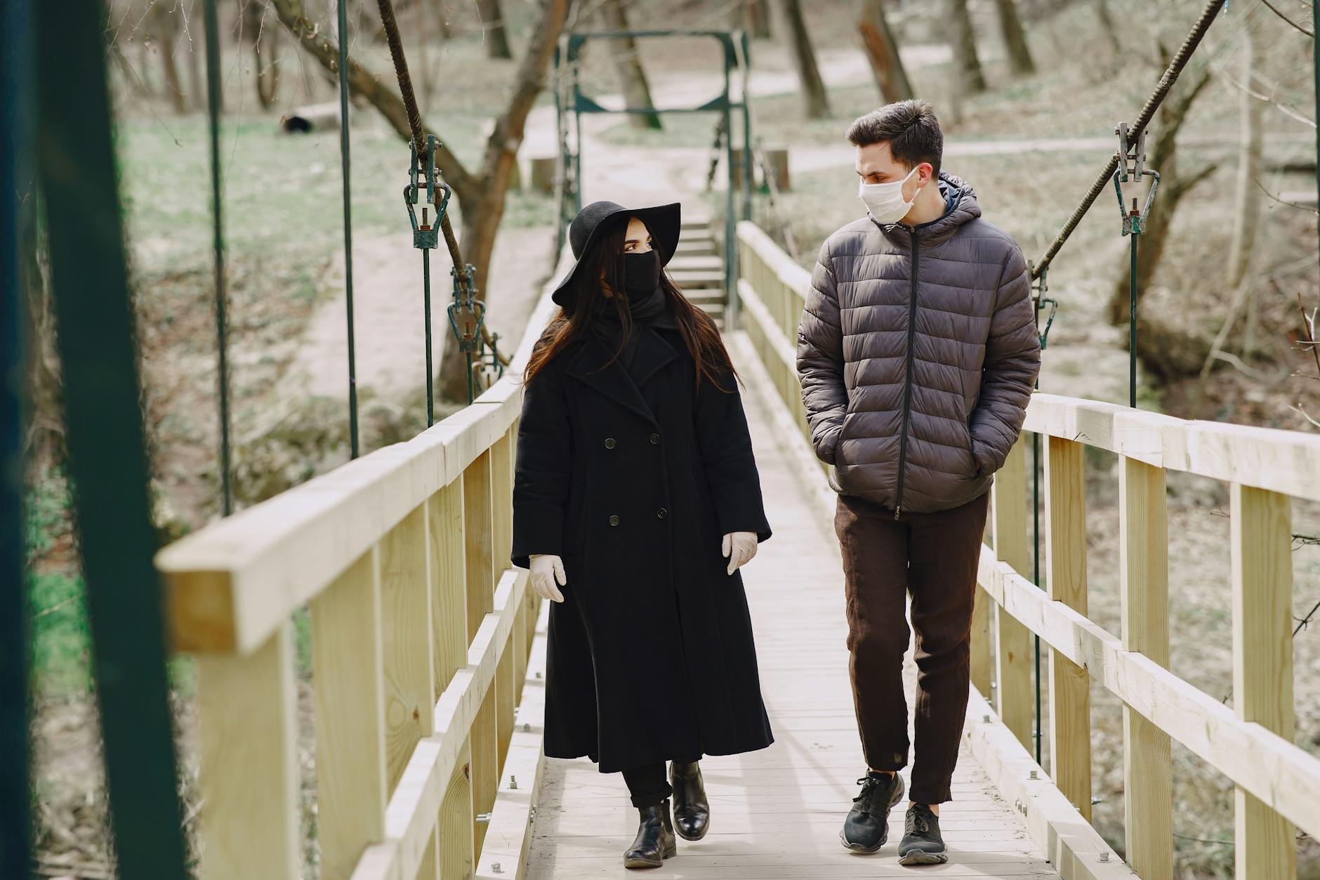A masked couple strolls on a wooden bridge in a park, observing social distancing guidelines. - cold weather couple communication