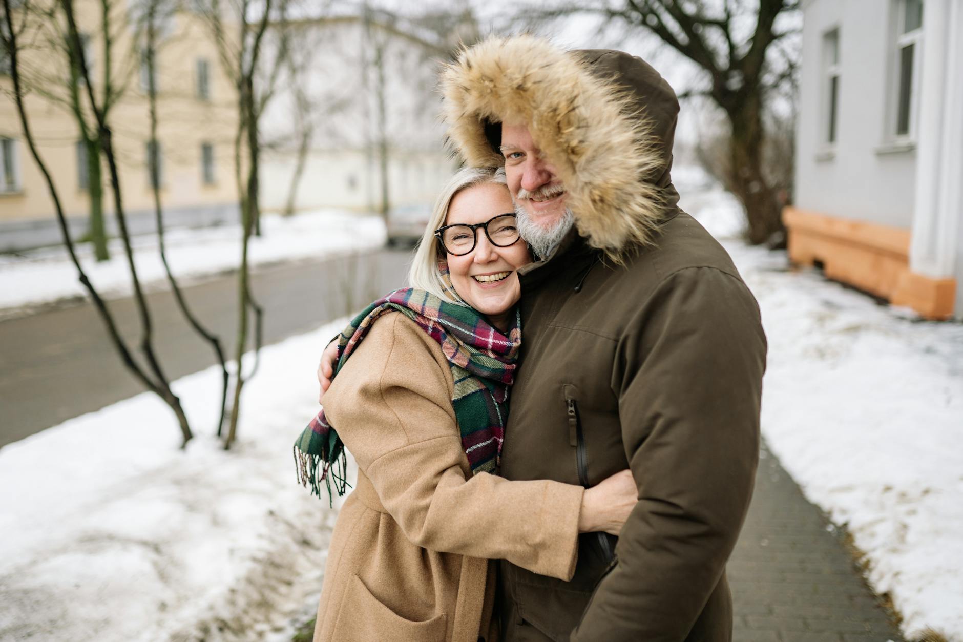 Happy senior couple in winter coats smiling and embracing outdoors on a snowy day. - cold weather couple communication