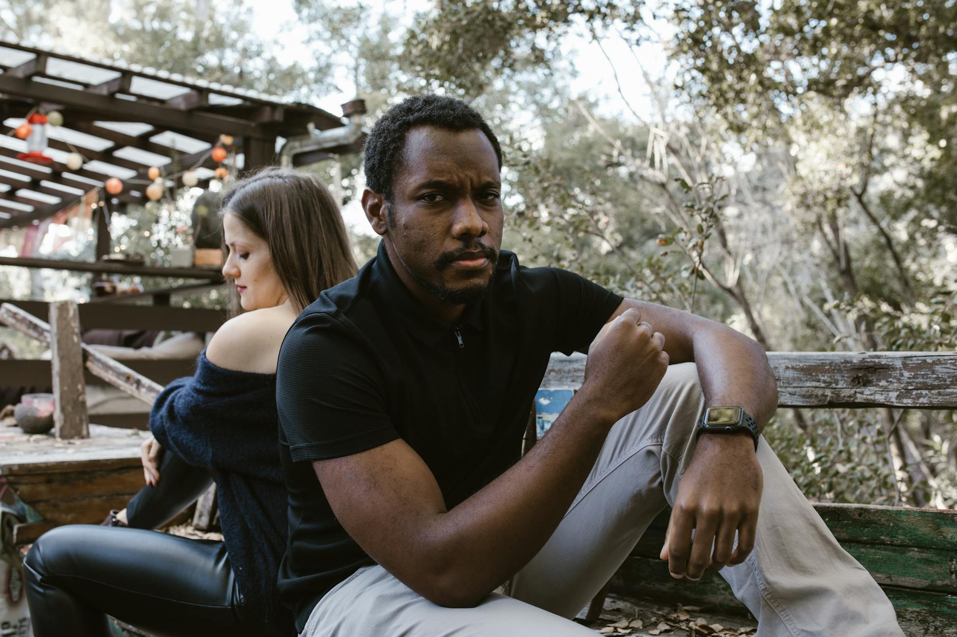 A thoughtful man sits outdoors, with a woman sitting distantly behind him. - how to cope with breakup