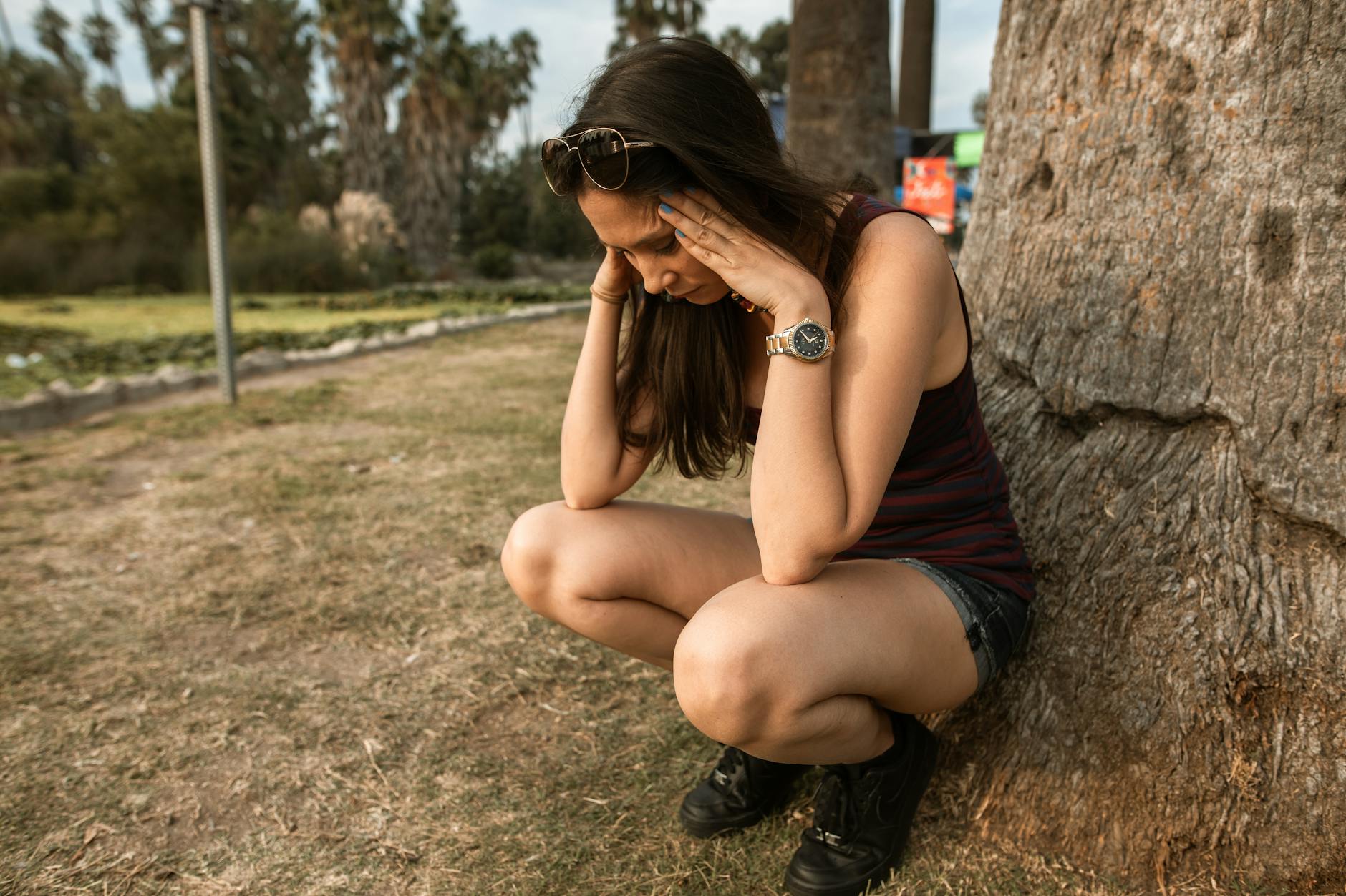 A woman kneels by a tree holding her head, expressing stress or anxiety in an outdoor park. - how to cope with breakup