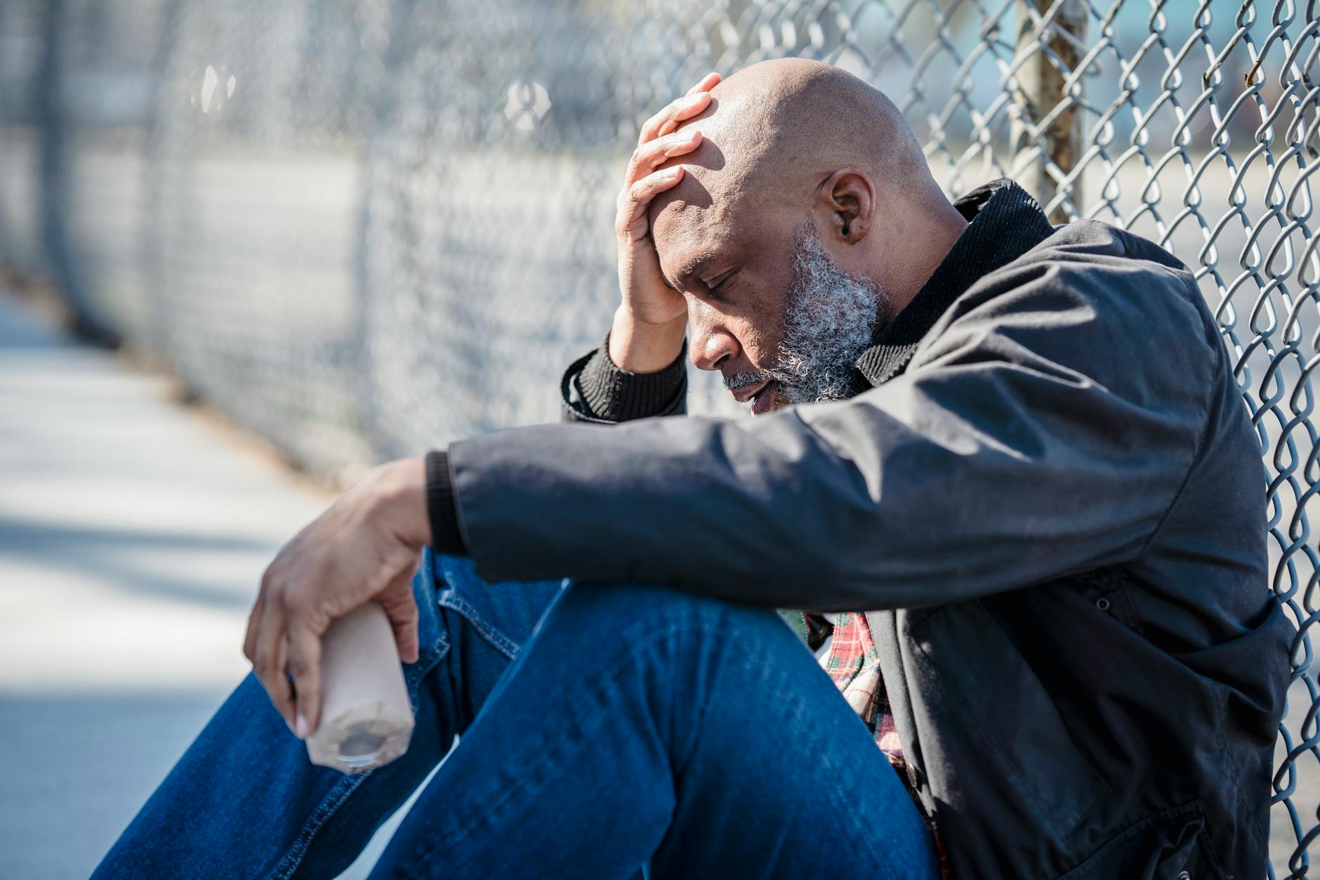 Pensive adult man with hand on head sitting by a fence, holding bottle. - coping skills for depression