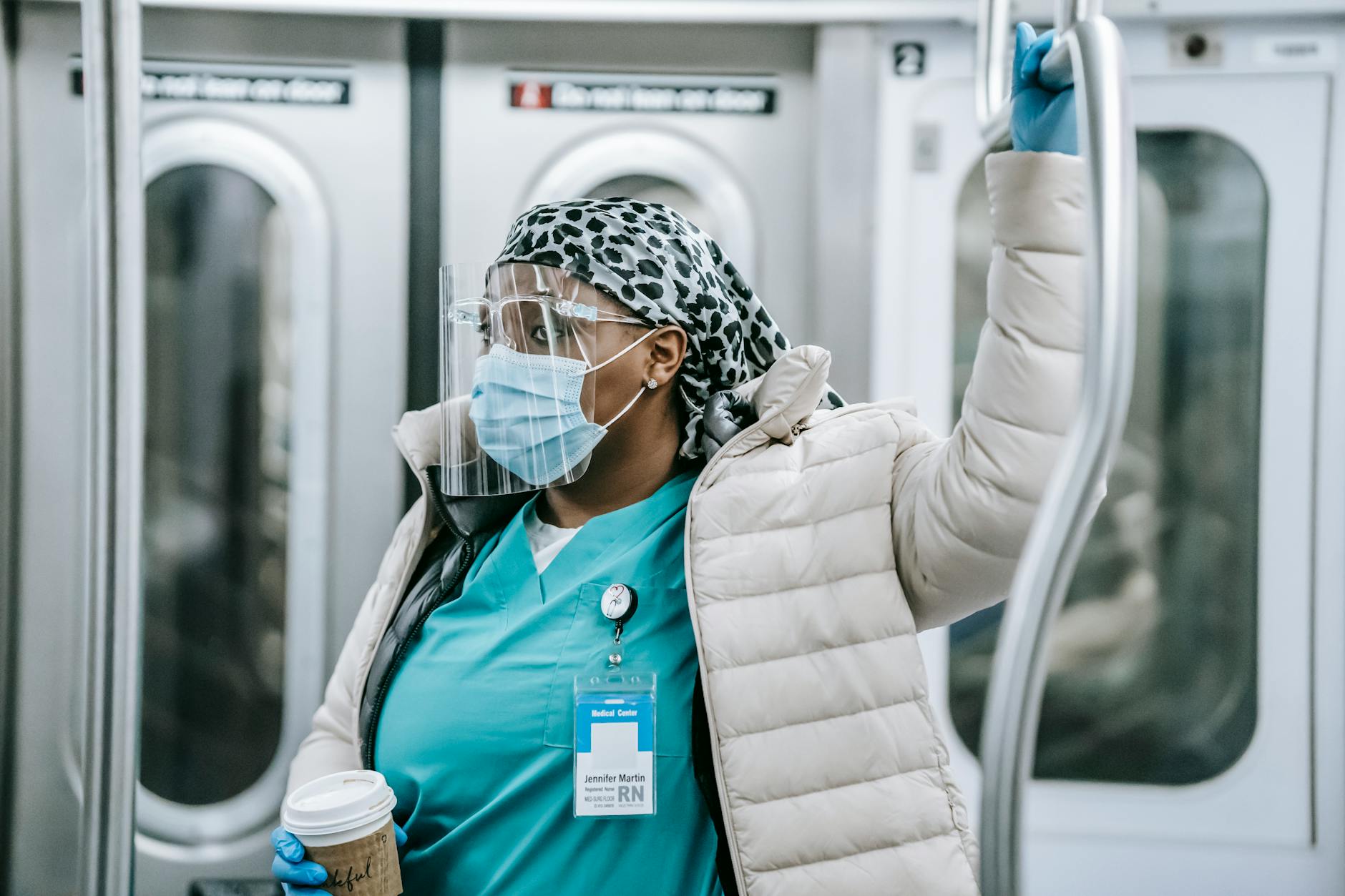Contemplative adult African American female doctor in uniform under warm clothes wearing protective face shield mask and gloves holding handle and riding metro train - coping with winter blues