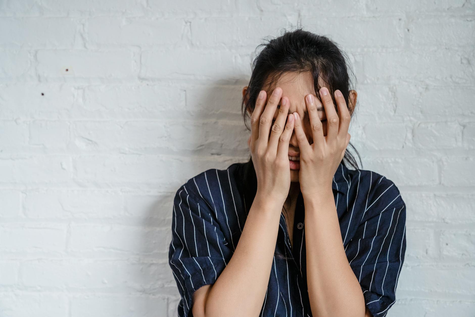 A young woman with hands covering face expresses stress against a white brick wall background. - coping with winter blues